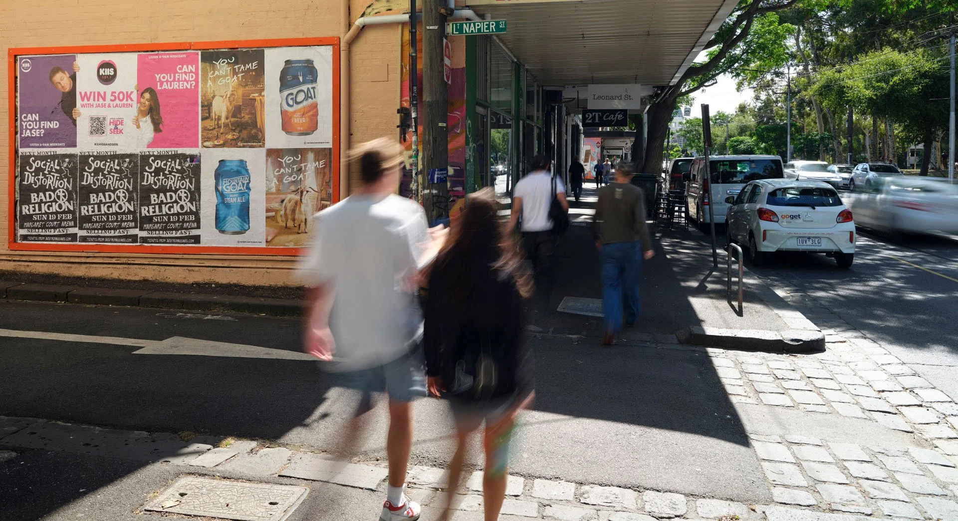 A busy city sidewalk with pedestrians walking past a storefront with posters on the wall, a street sign reading L' Napier St, and a row of parked cars along the curb. Trees line the street, casting shadows on the pavement.