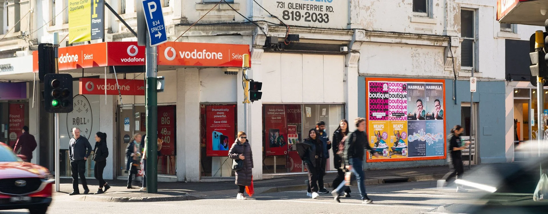 Street scene in an urban area shows pedestrians crossing the street near a storefront with Vodafone signage. There are traffic lights, a bus, and various advertisements on the building walls.