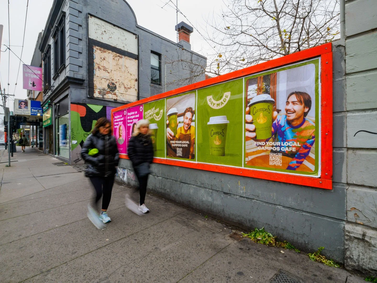 Street scene with two women walking past a sidewalk with colorful advertisements for Campos Café on the building wall. The ads feature a smiling man holding a coffee cup, and include QR codes and text promoting finding local Campos Café locations.