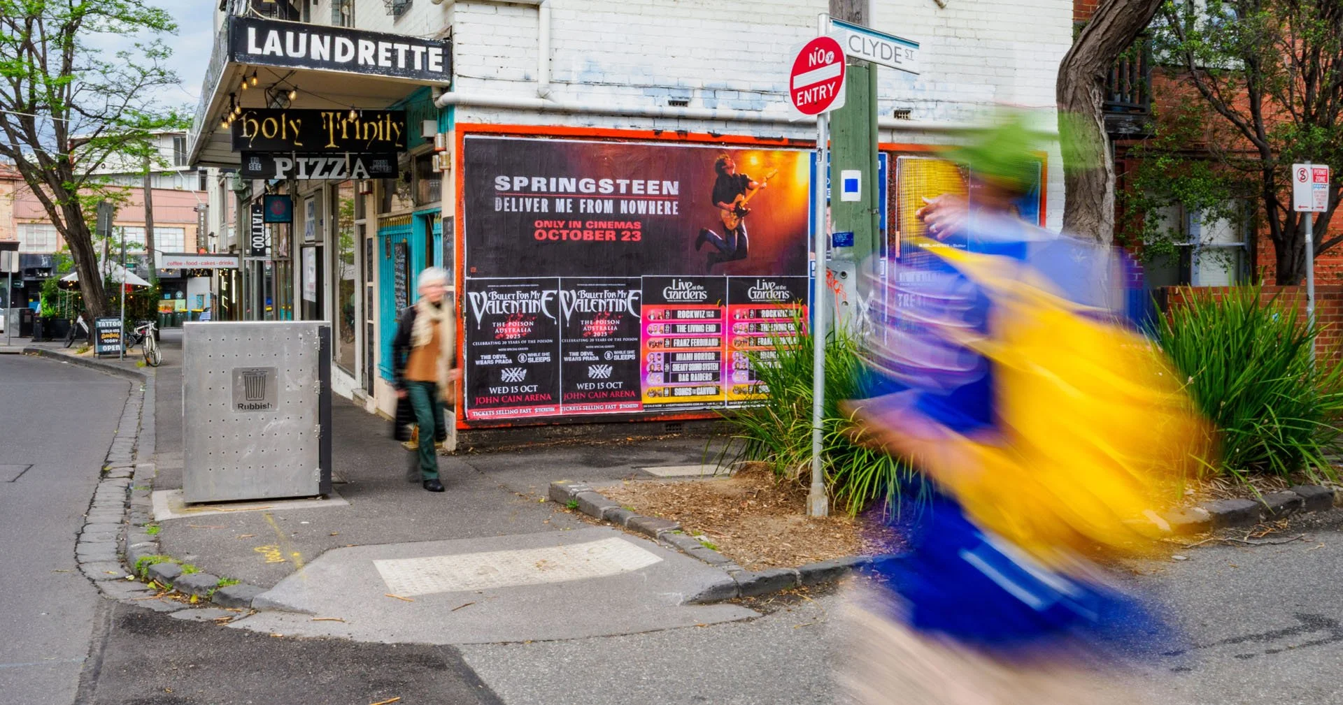 Corner of a street with a poster advertising Springsteen's concert and a woman walking past. Blurred figure on a scooter moving quickly in the foreground. Buildings and trees surround the scene.