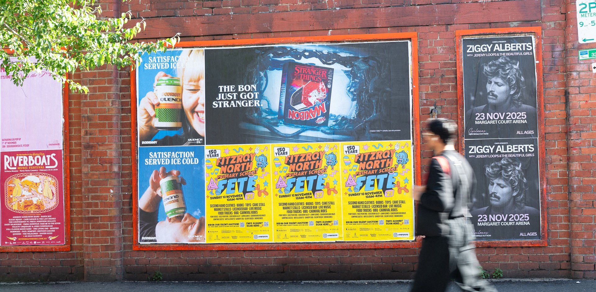 Brick wall featuring various posters including an ice cream ad, a Stranger Things magazine cover, and an event poster for Fitzroy North Primary School's fete. A person in motion walks past on the sidewalk.