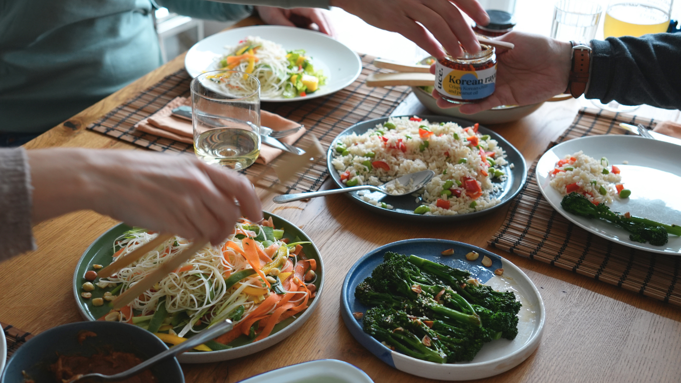 Family sharing a Korean-inspired meal with rice, noodles, vegetables, and sauce at a wooden table.