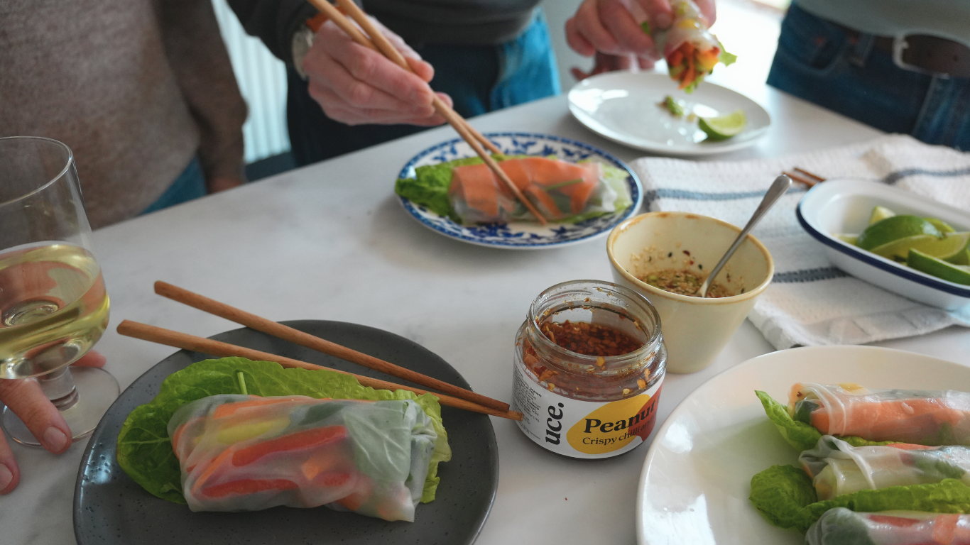 Three people eat summer rolls at a kitchen counter, with a jar of uce peanut rayu in the middle of the shot