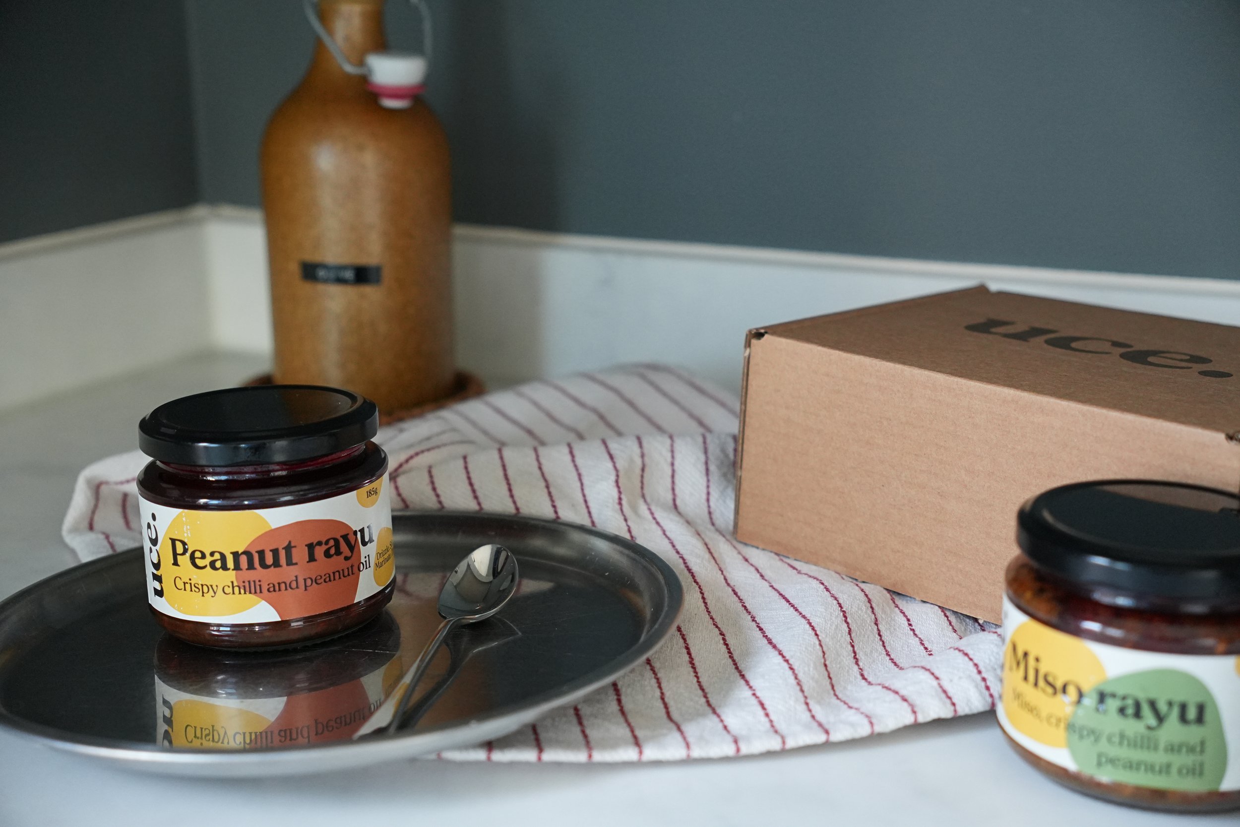 In the foreground is a jar of peanut rayu on a silver tray along with a teaspoon. Out of focus is an 'uce' packaging box and a jar of miso rayu