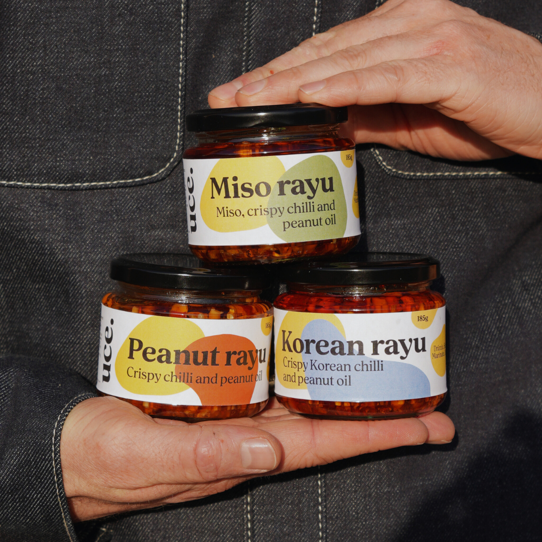 A person holding three jars of uce: Miso on top, peanut bottom left and Korean bottom right. They have black lids and colourful labels. The person is wearing a dark denim shirt.