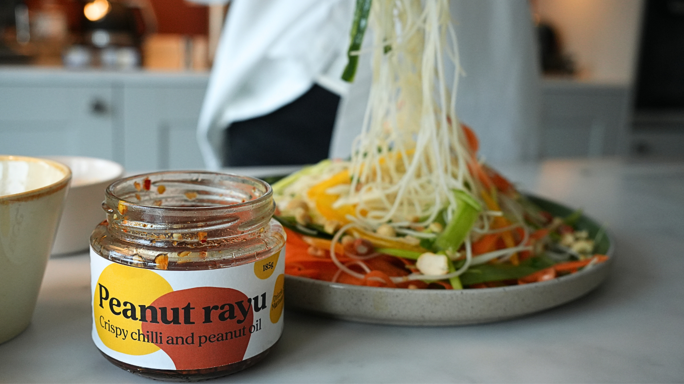 A jar of uce peanut rayu on a kitchen counter in front of a plate of noodles behind and softly out of focus