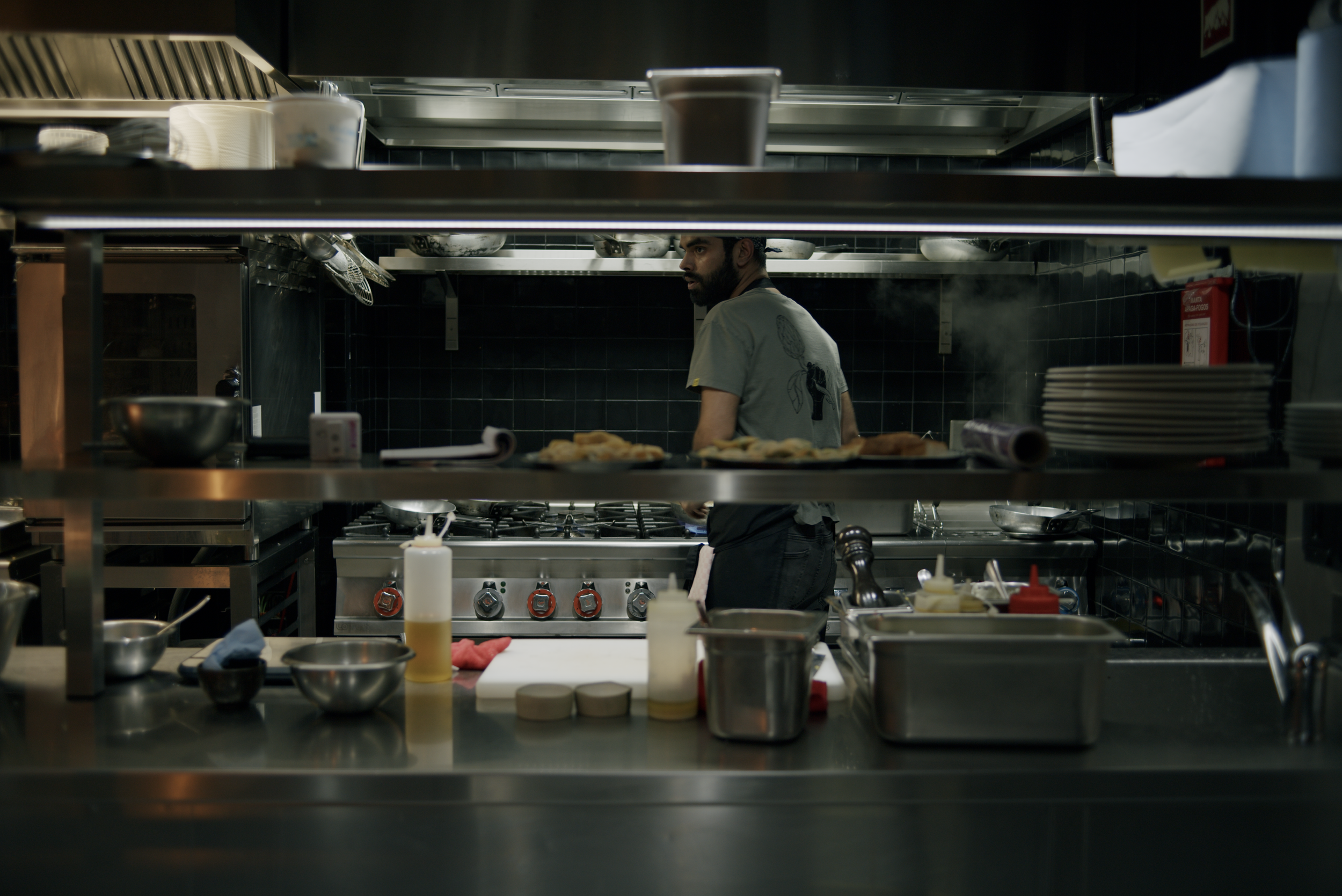 A male chef working in a professional kitchen, seen through a gap in the kitchen shelf, with pots, pans, and kitchen utensils around.