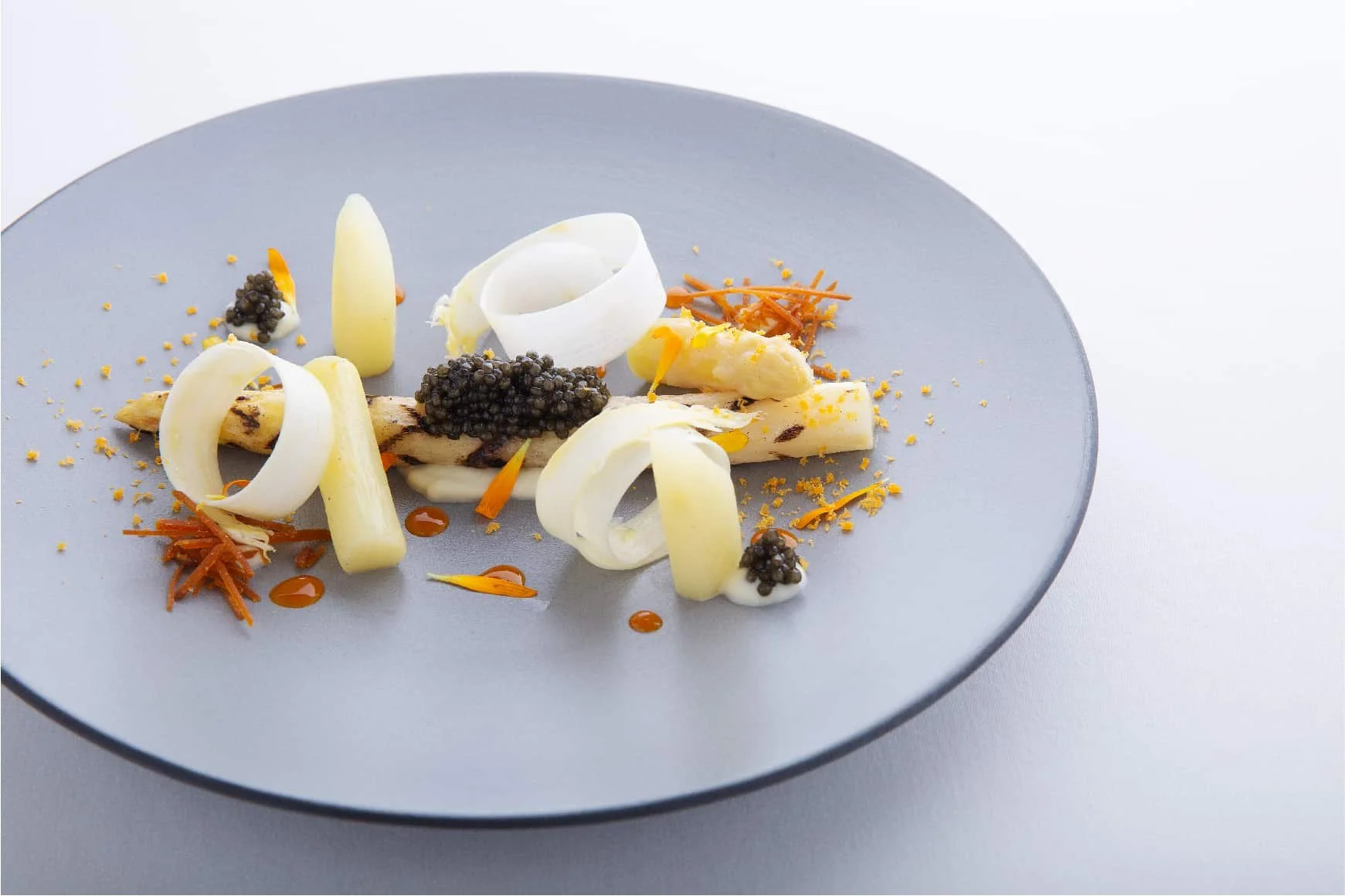 Artfully plated dish with curled white asparagus ribbons, black caviar, shredded bottarga, on a gray plate.