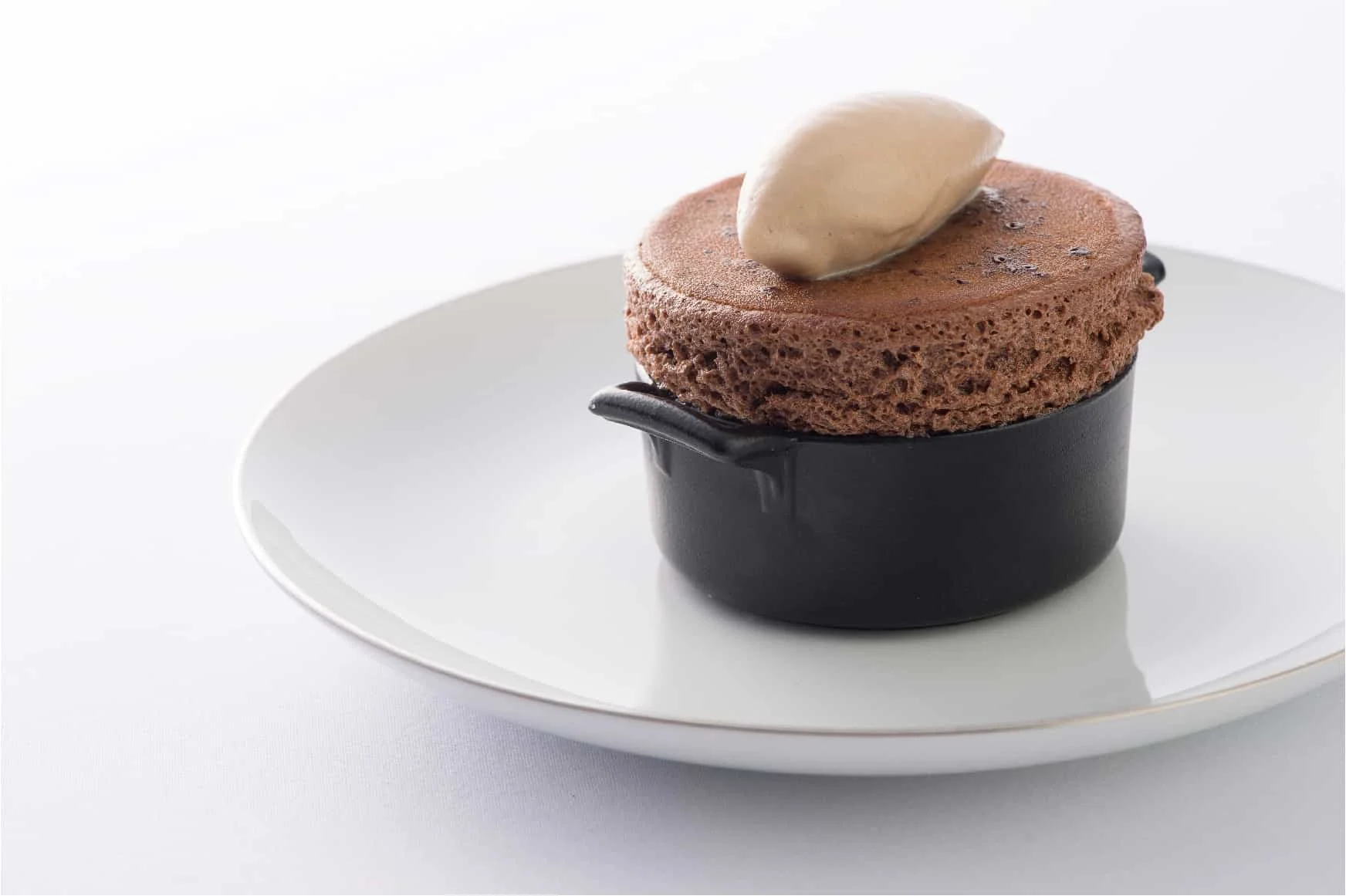 Chocolate souffle dessert in a black ramekin with a dollop of hazelnut ice cream on top, served on a white plate.