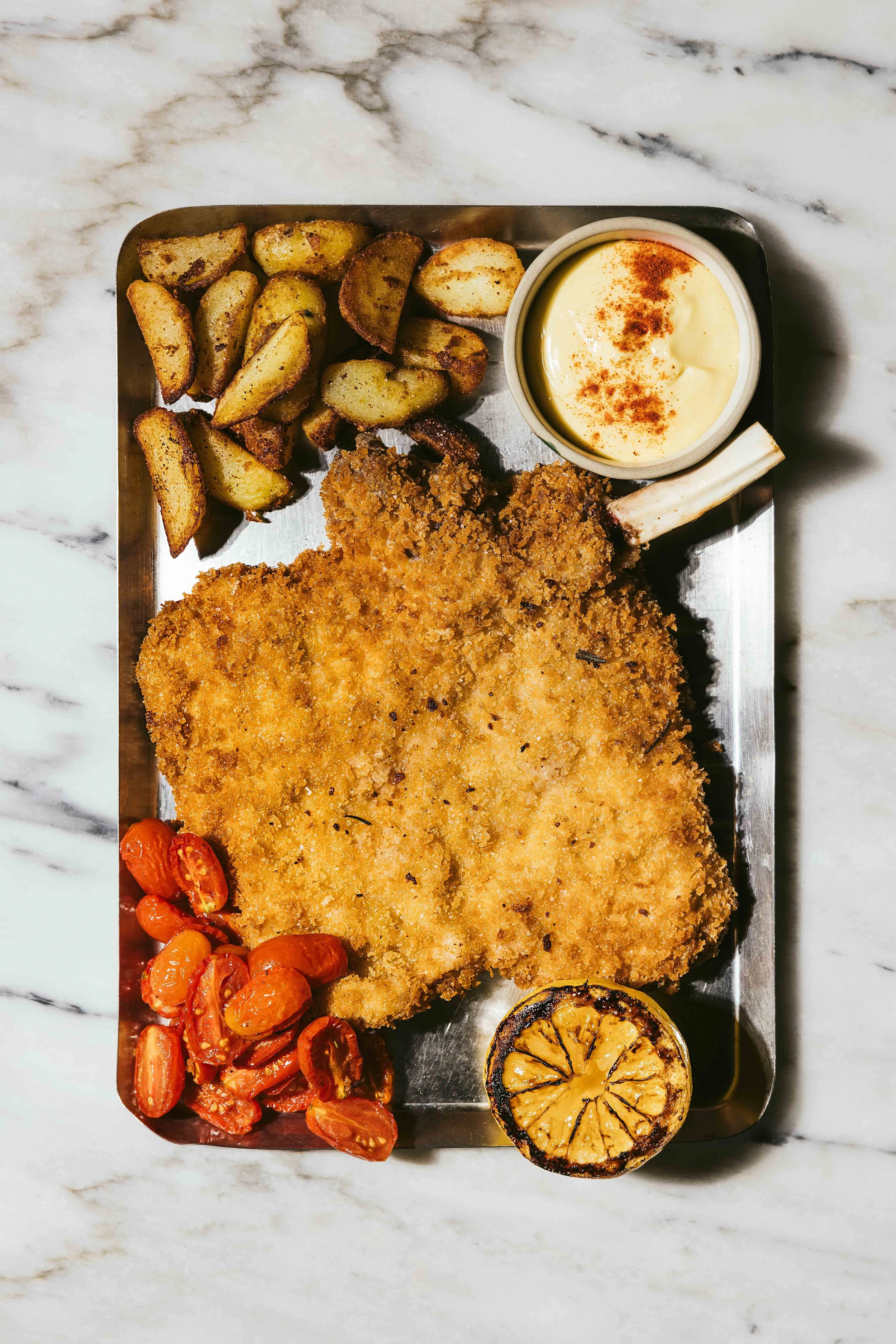 A metal tray with breaded, fried veal chop, roasted potatoes, cherry tomatoes, a bowl of creamy mayonnaise with paprika, and a grilled lemon half.