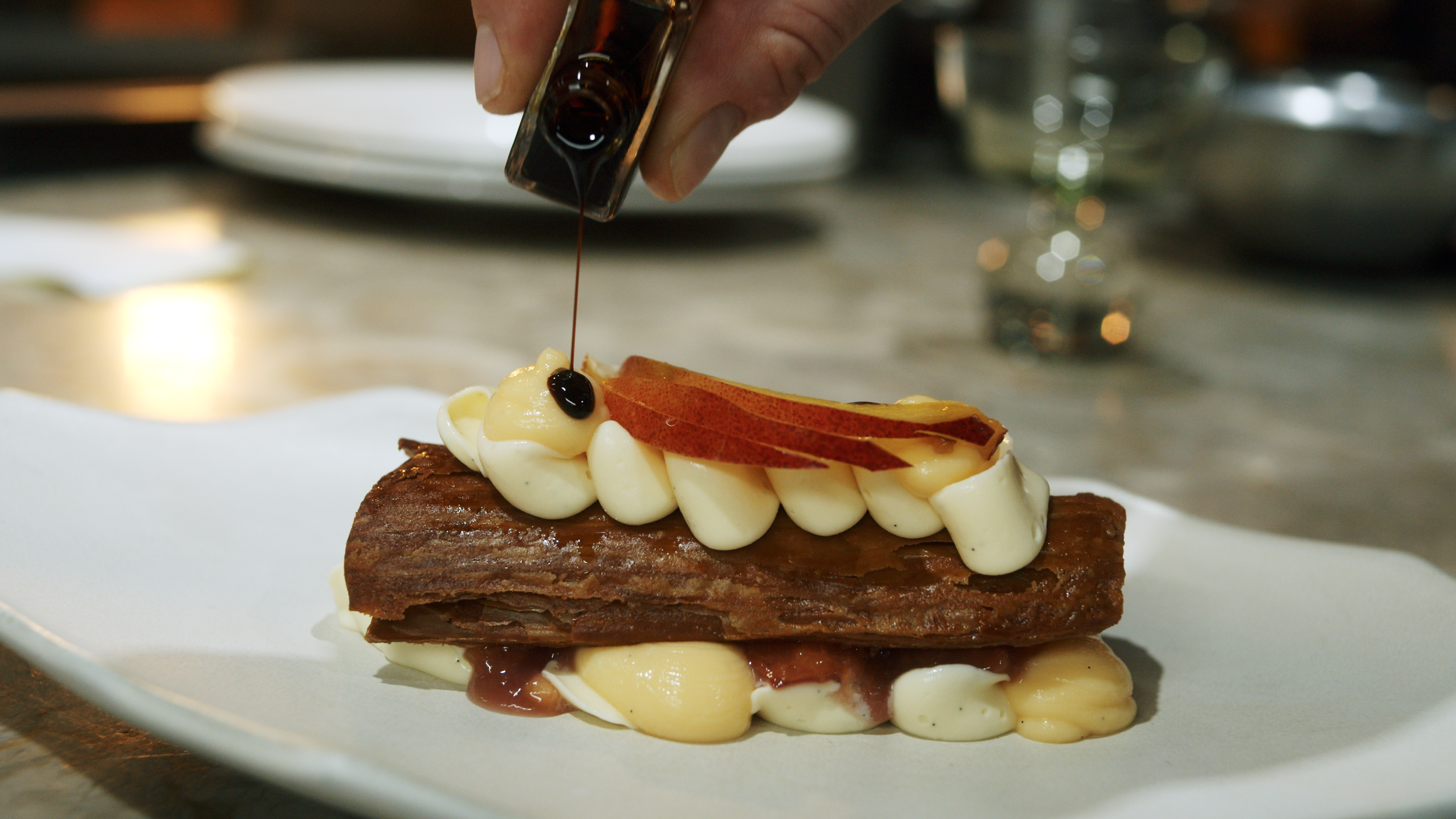 A millefuille dessert plate with pastry cream, topped with white frosting, peaches slice, and a drizzle of dark aceto balsamico tradizionale di Modena, being added by a hand.