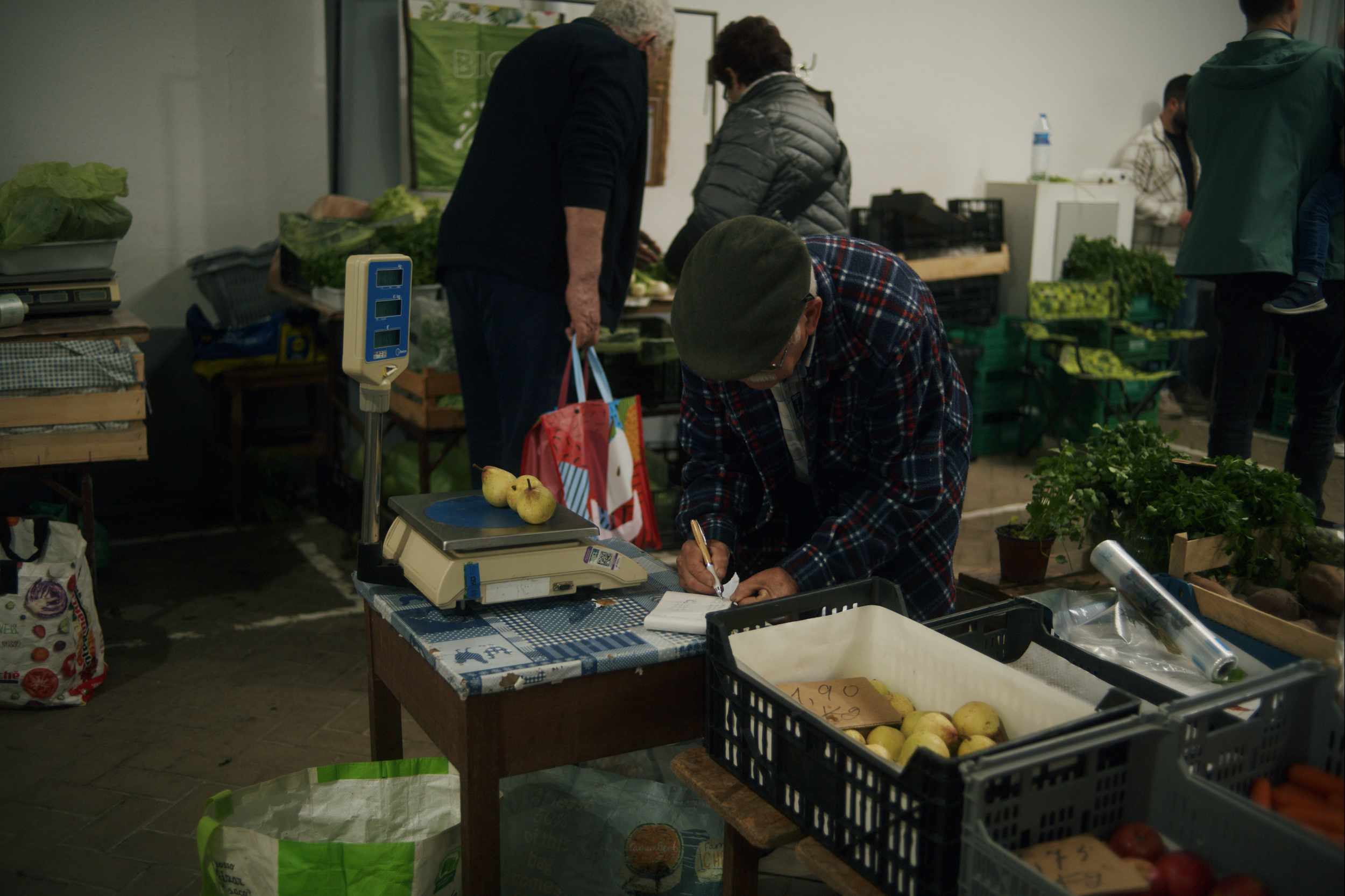 People shopping at a farmers' market, examining and writing on a notepad at a stall with apples and vegetables.