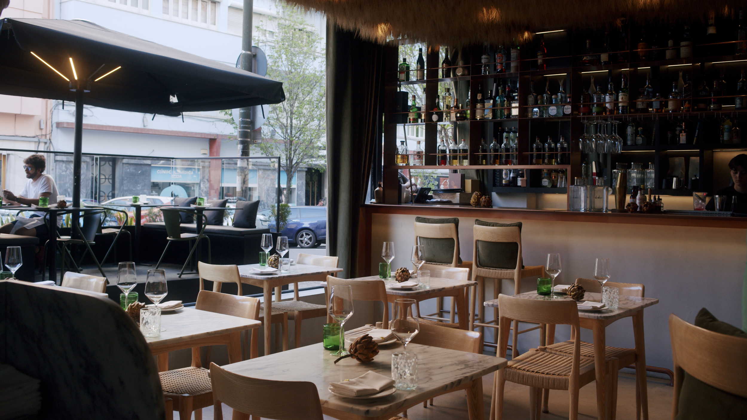 Interior of a restaurant with wooden tables set with wine glasses, green glass vases, and napkins. Outside view through large windows shows a man seated at a table under an umbrella. The bar area has shelves filled with bottles and glasses.