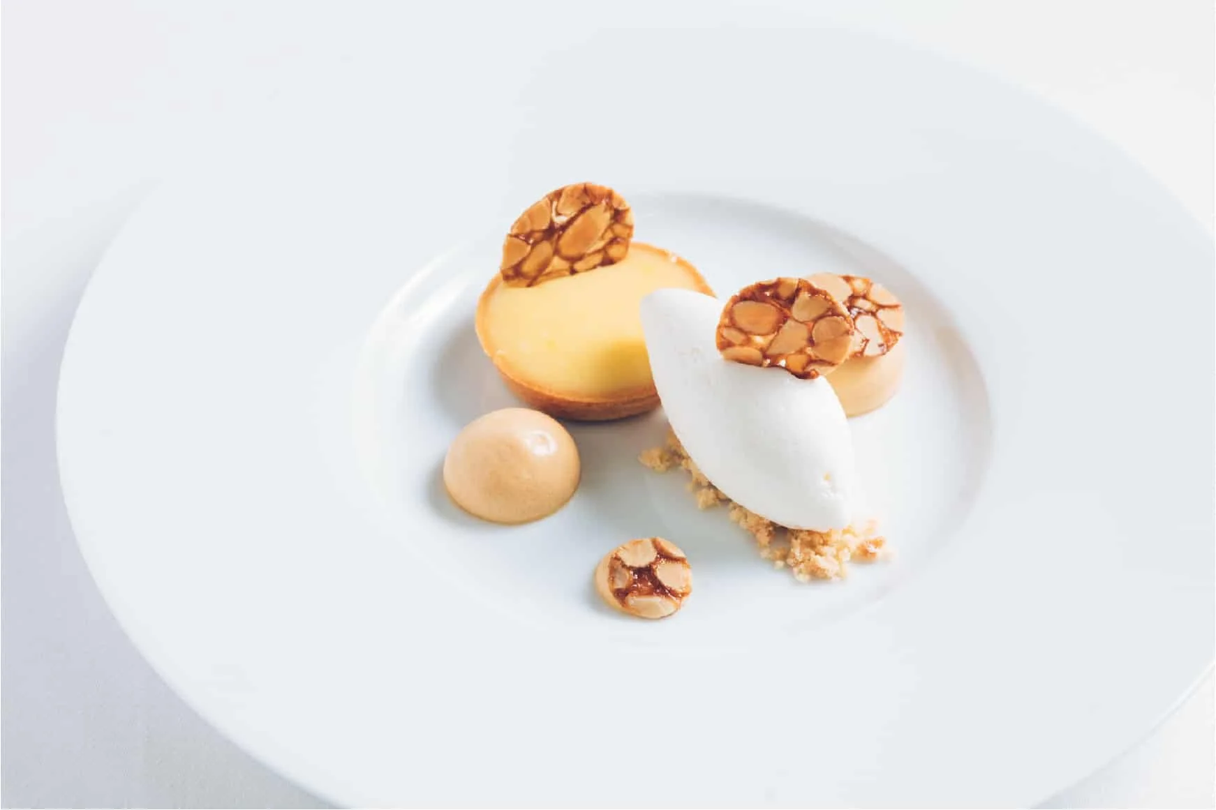 Elegant dessert plate featuring a halved pastry with lemon creamy filling, a dollop of smooth caramel sauce, a quenelle of latte ice cream, and several caramelized almond toppings, arranged on a white round plate.