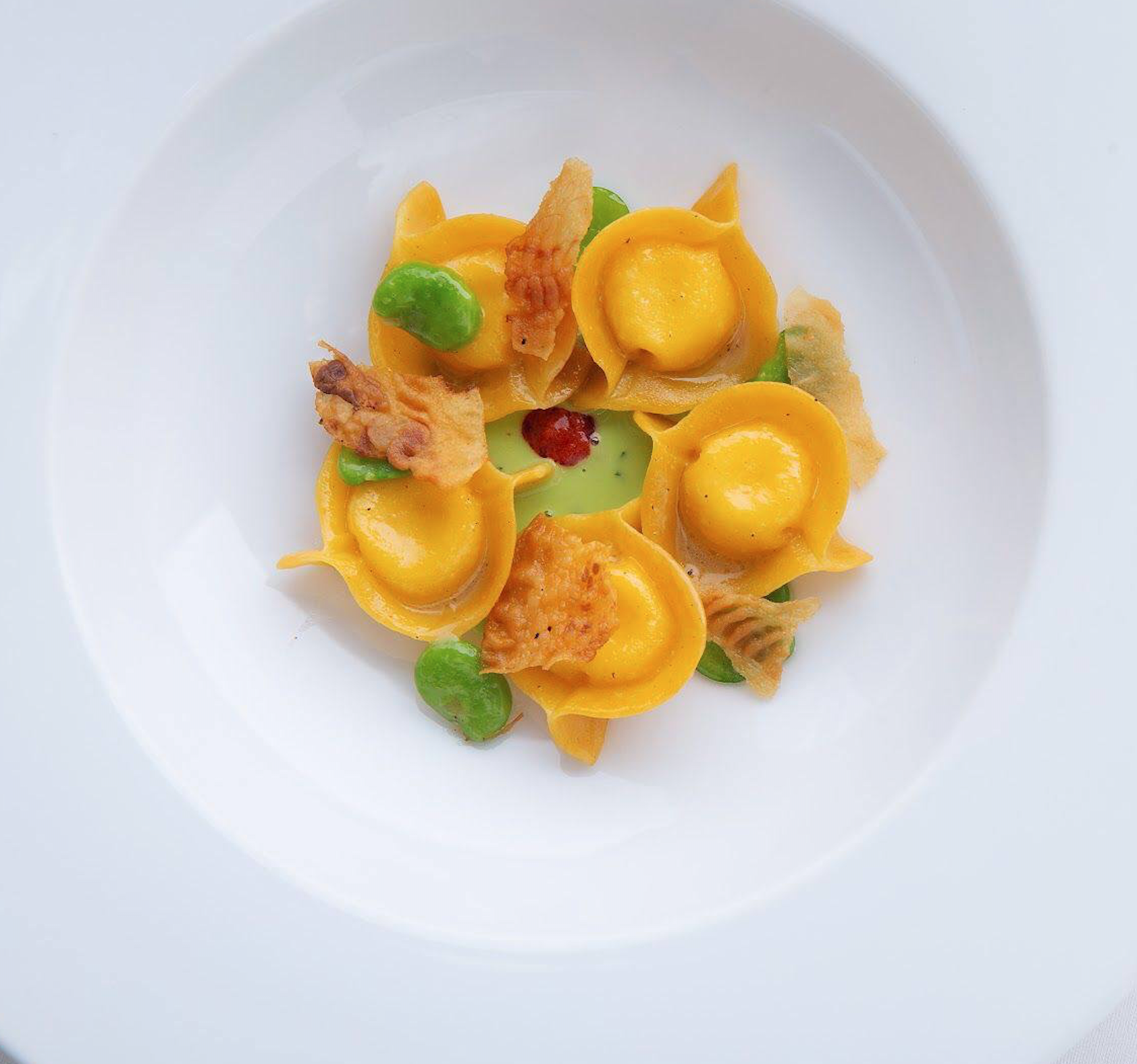 Close-up of yellow tortellini pasta with green peas, crispy guanciale pieces, and red nduja, served on a white plate.