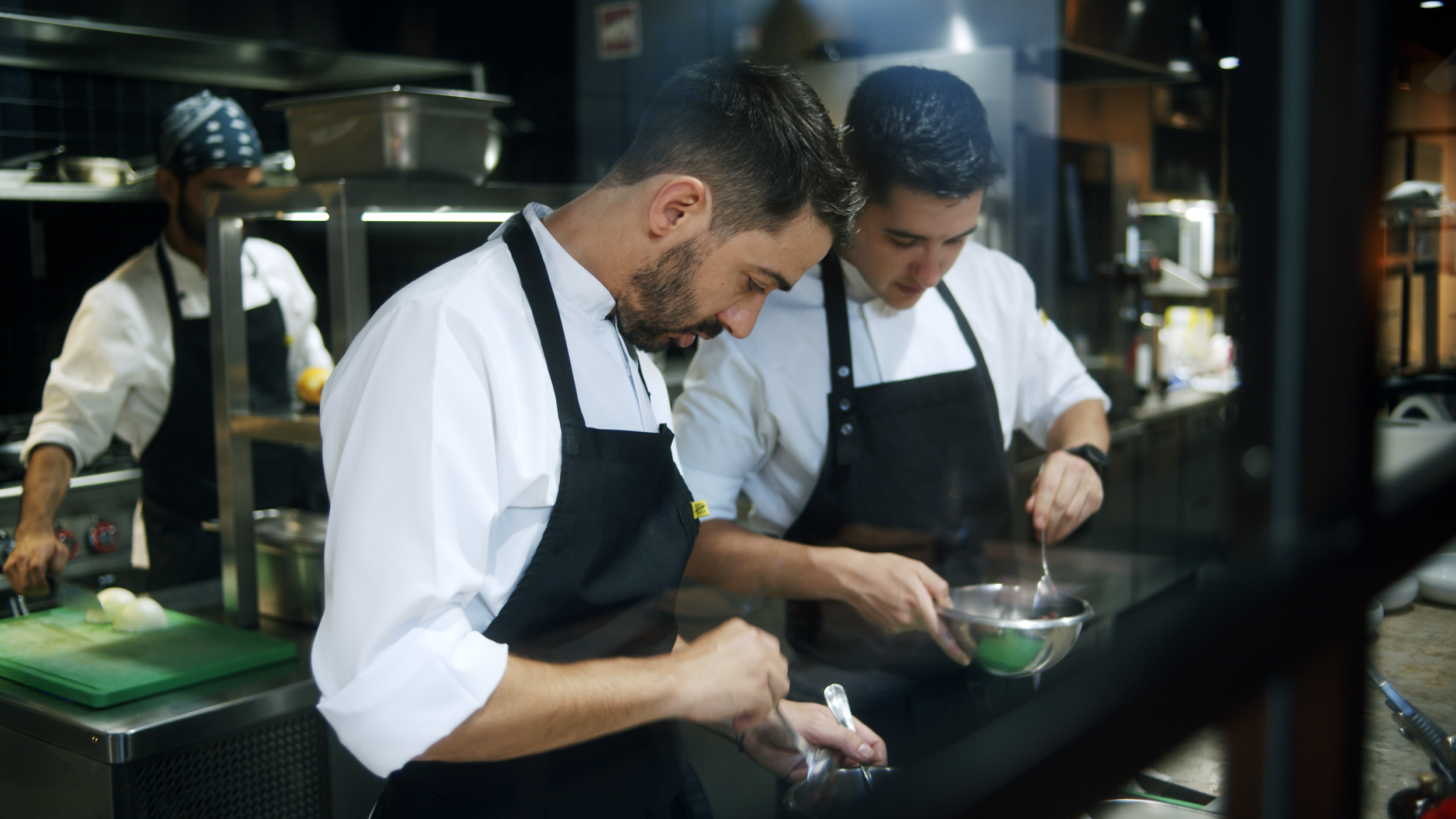 Two chefs in white uniforms and black aprons preparing food in a professional kitchen, with another chef working in the background.