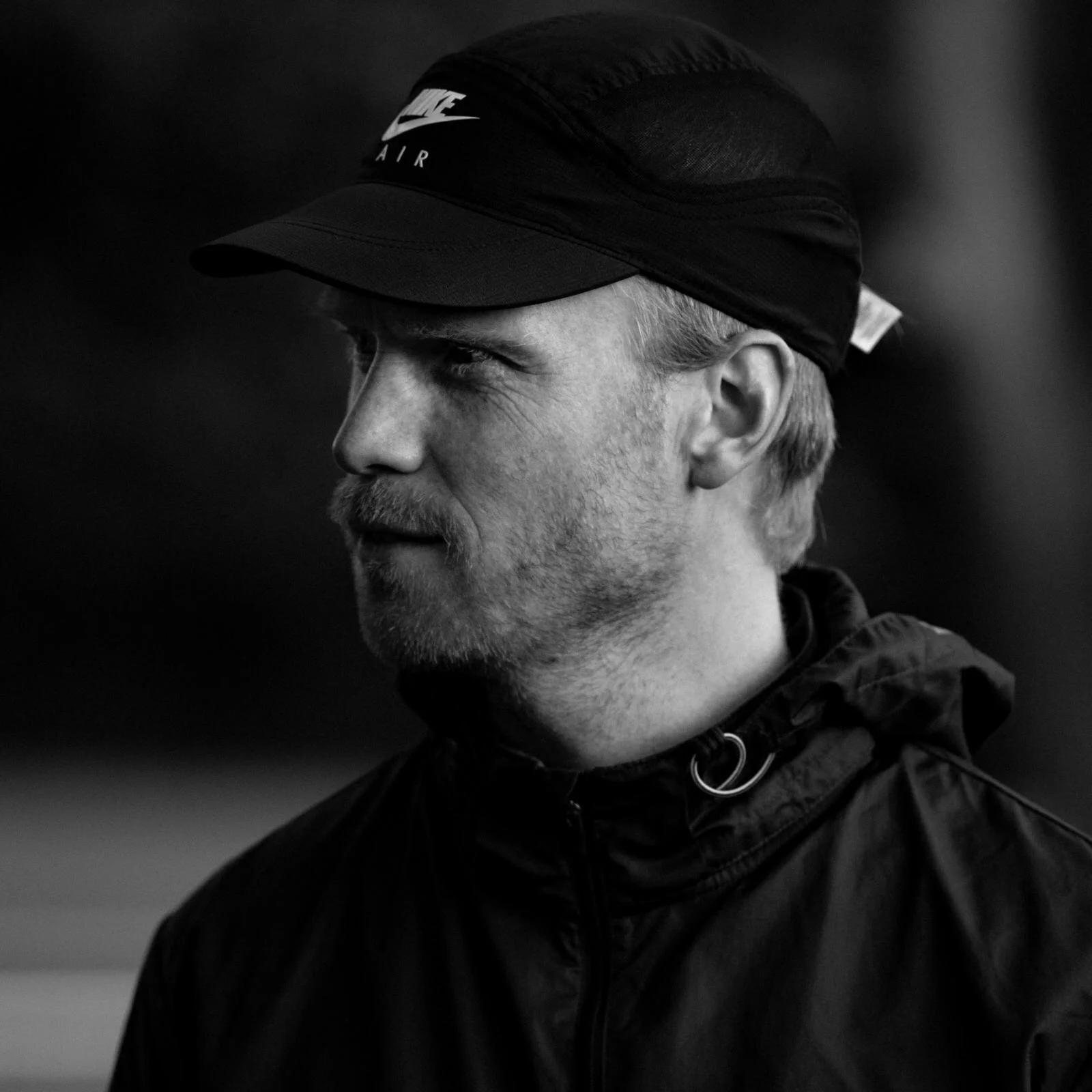 Een zwart-witportret van een man die een Nike Air-pet en een donkere jas draagt, en met een geconcentreerde blik naar links kijkt.