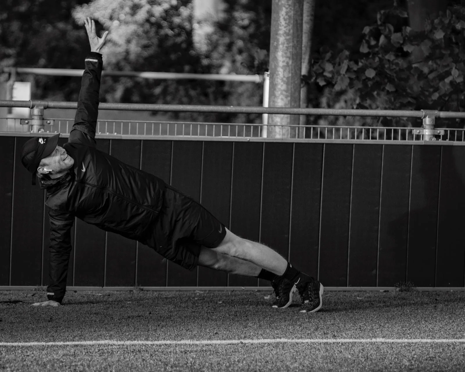 Een man die een eenarmige side plank doet op een atletiekbaan, gekleed in sportkleding en met een pet op.