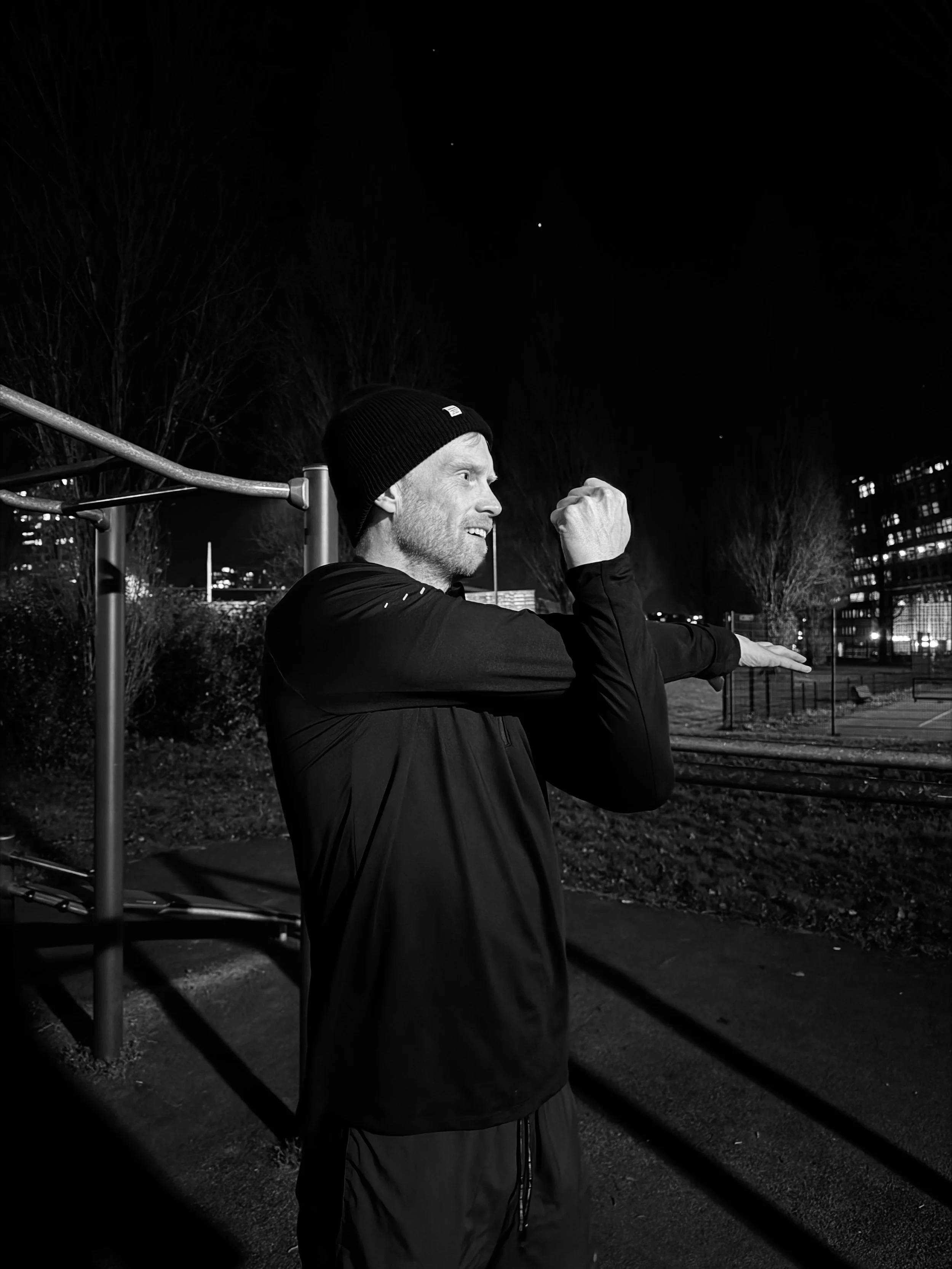 Een man in sportkleding en een muts op die ’s avonds buiten een stretch oefening doet.