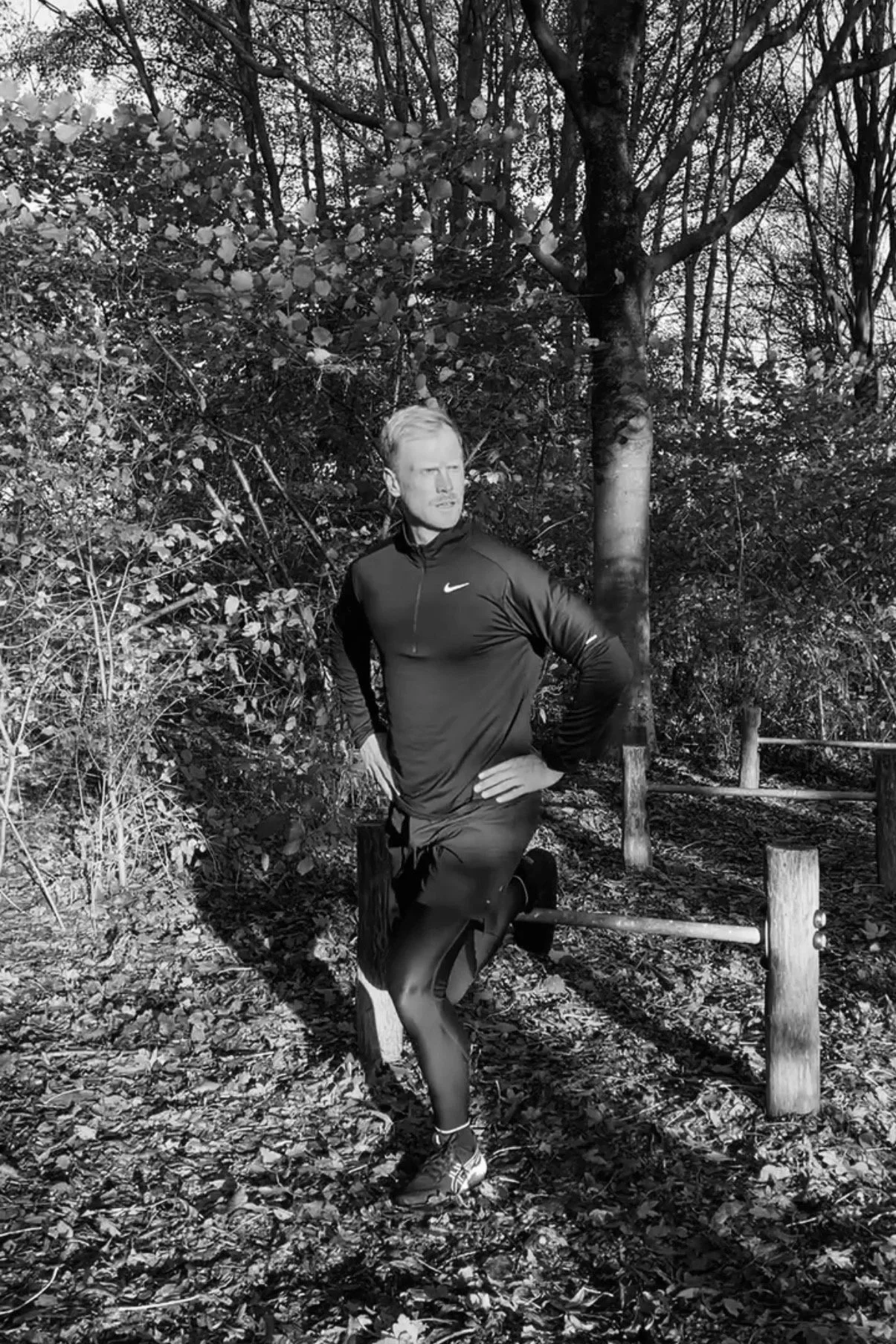 Een man in sportkleding die buiten tussen bomen en begroeiing staat en een bulgarian split squat doet met z'n handen in z'n zij.