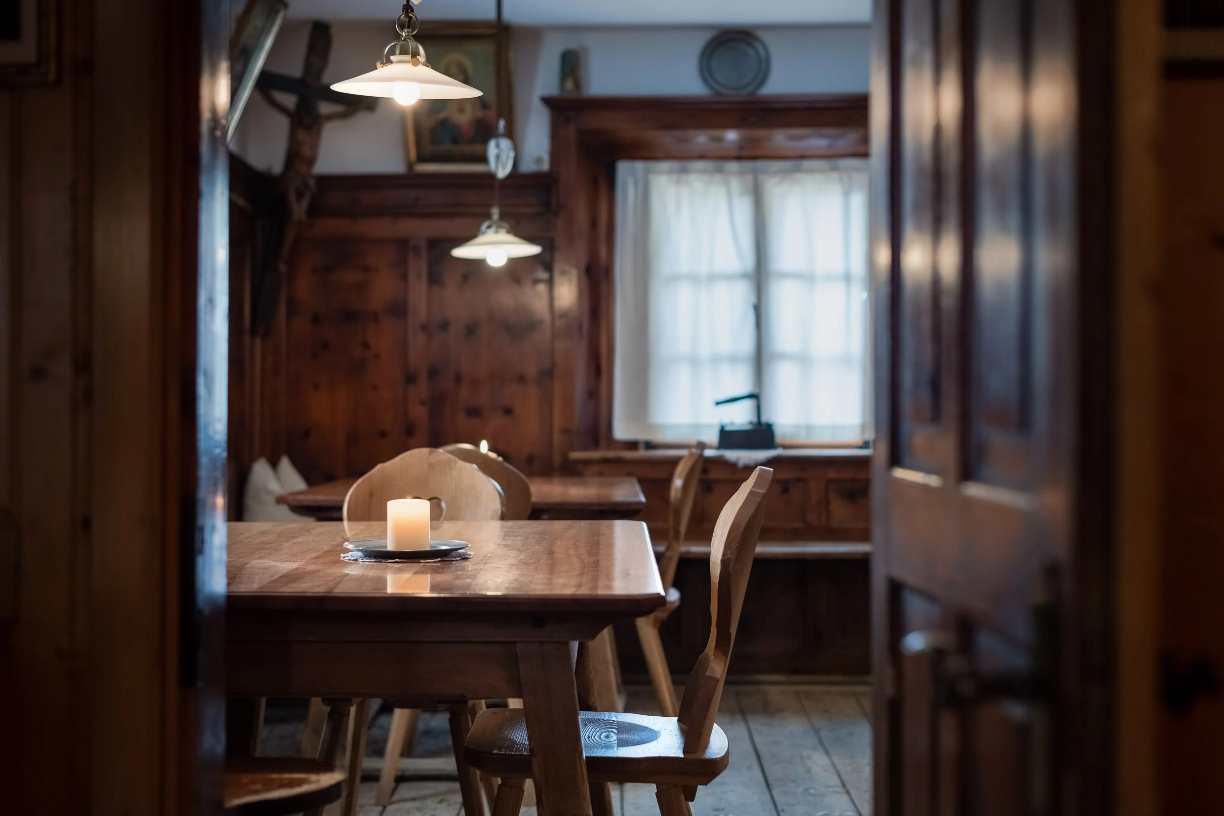 A cozy wooden dining room with a table set with a lit candle, surrounded by wooden chairs, and illuminated by hanging pendant lights. There's a window with white curtains, and a corner with a desk and a wall-mounted iron.