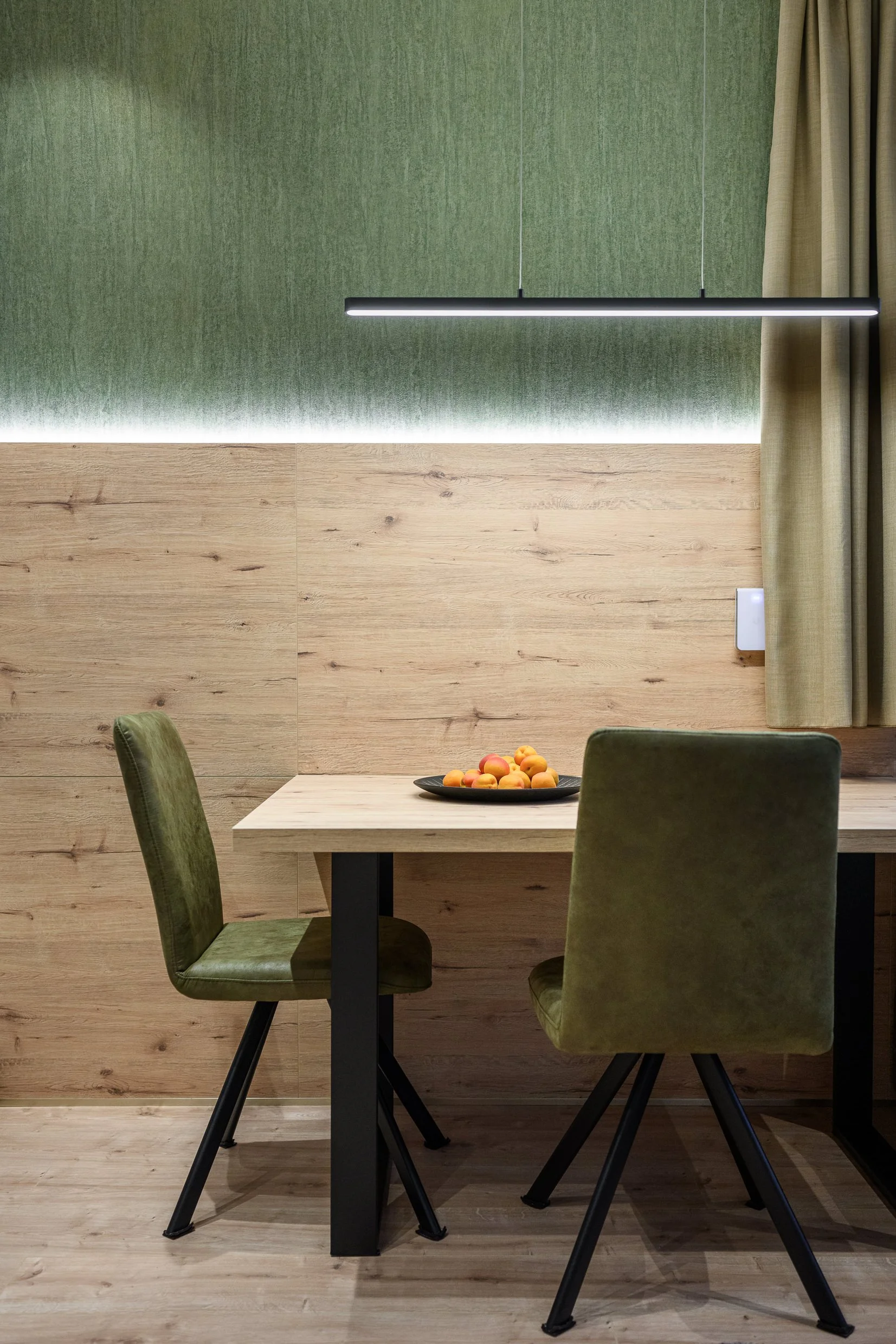 A wooden dining table with two green velvet chairs, a black plate with peaches, and a green textured wall with a modern horizontal hanging light fixture.