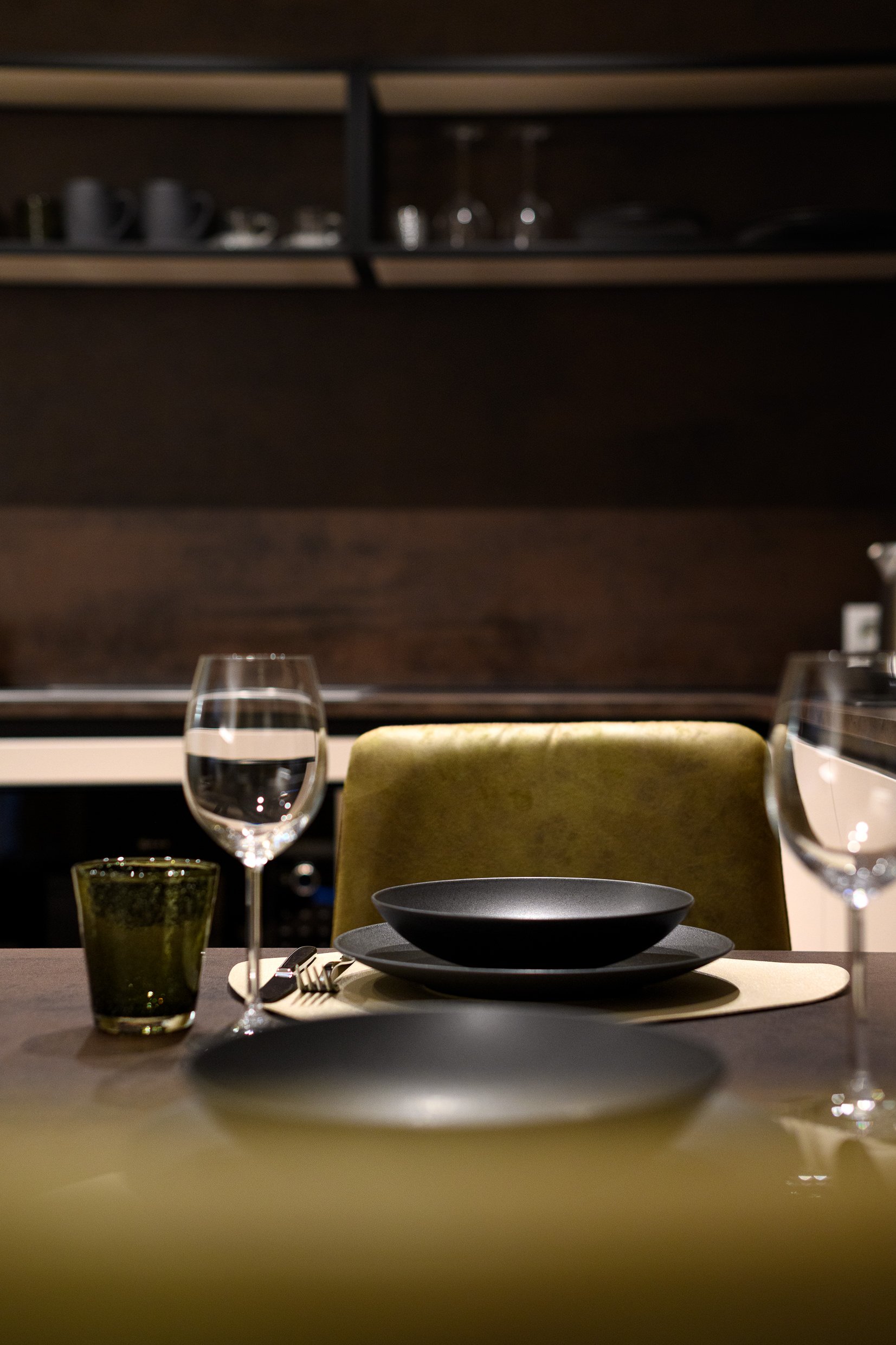 Empty restaurant table with black bowl, water glass, wine glass, and green tumbler, set against a dark wooden background with a green chair.