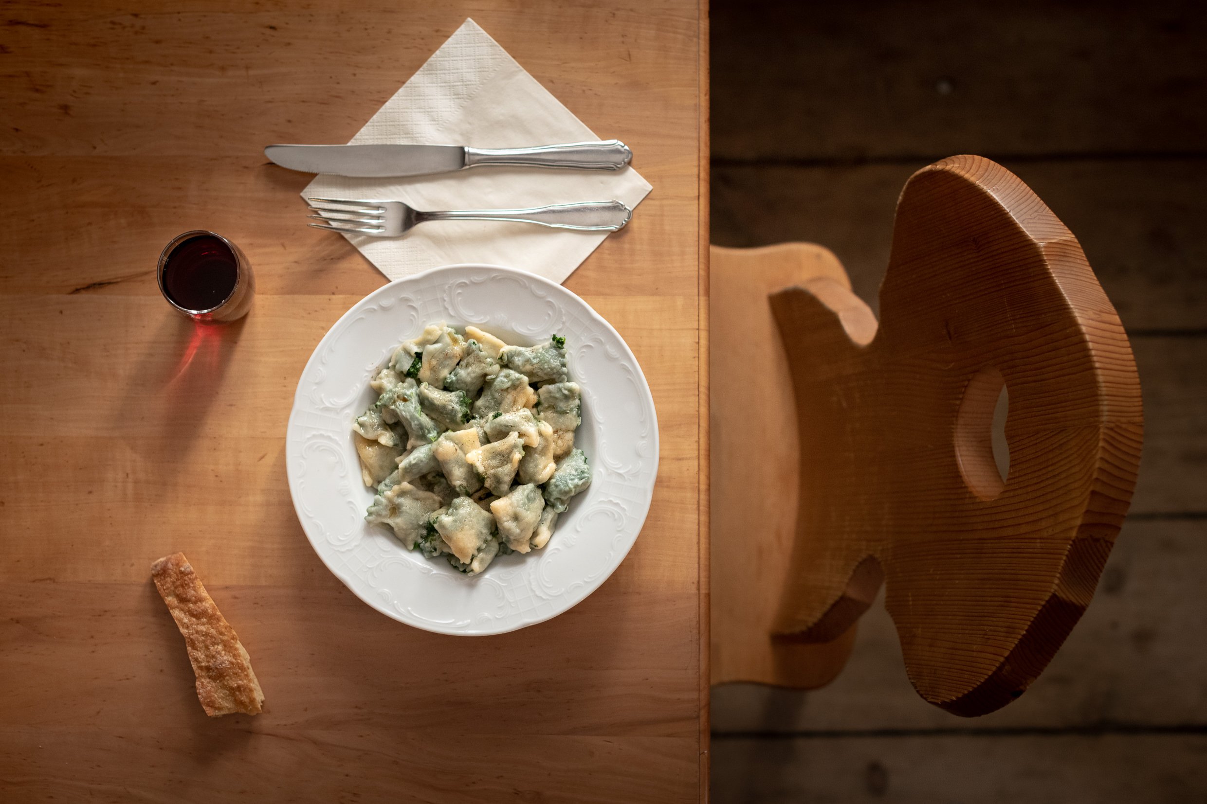 A top view of a wooden table with a plate of green-tinted tortellini pasta, a glass of red wine, a single breadstick, and a fork and knife on a napkin, with a wooden chair to the right.