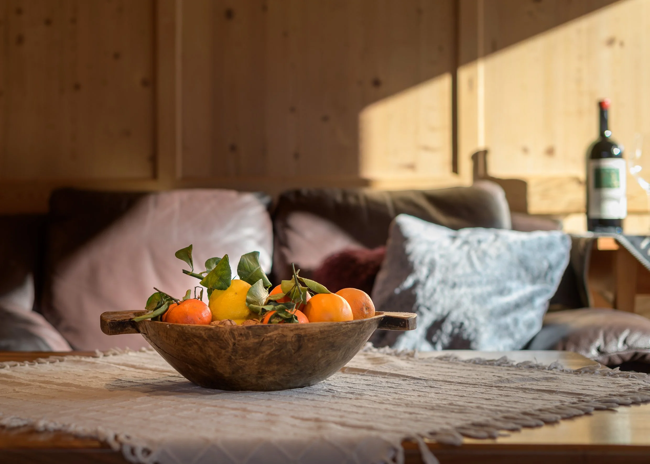 A wooden bowl with oranges, lemons, and green leaves on a woven tablecloth, with pillows and a bottle of wine in the background.