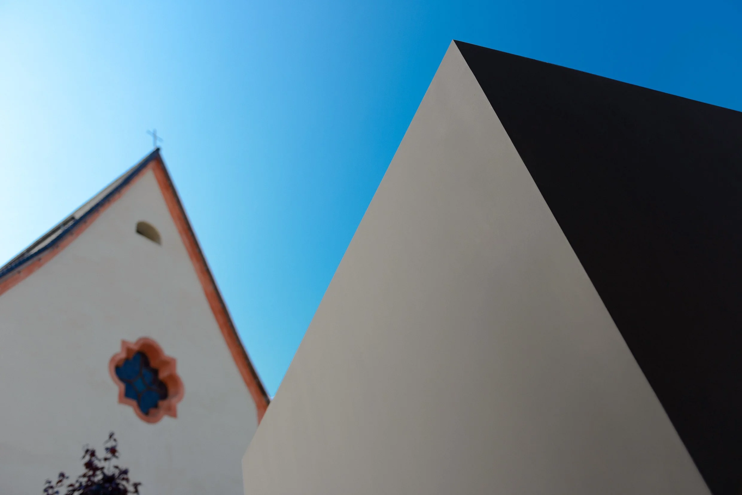 View of a church steeple with a cross and a rose window on a white wall, with adjacent sharp-edged modern building, against a clear blue sky.