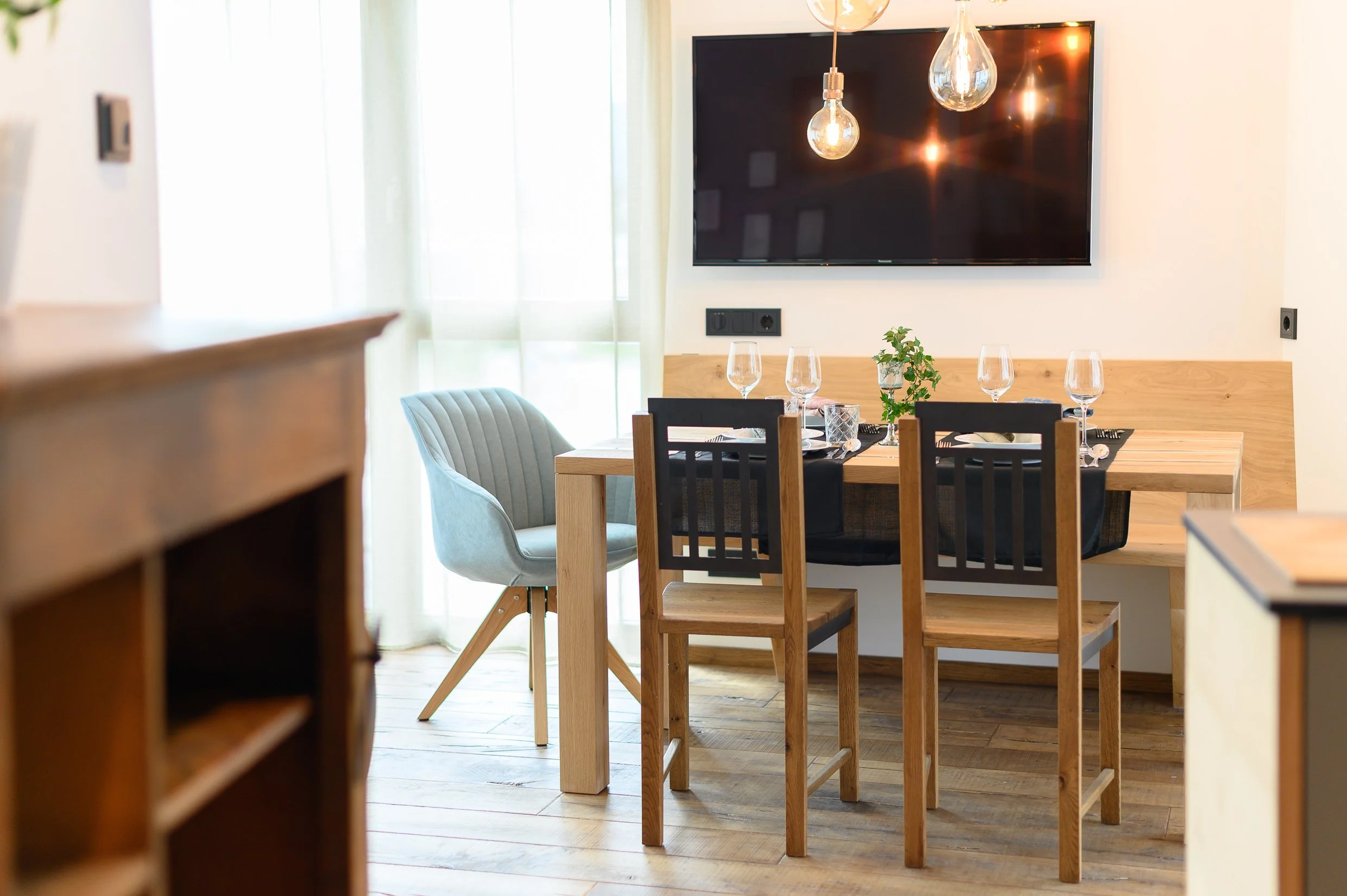Modern dining room with a wooden table set for four, three black and one gray chairs, hanging pendant lights, a large wall-mounted TV, and natural light coming through sheer curtains.