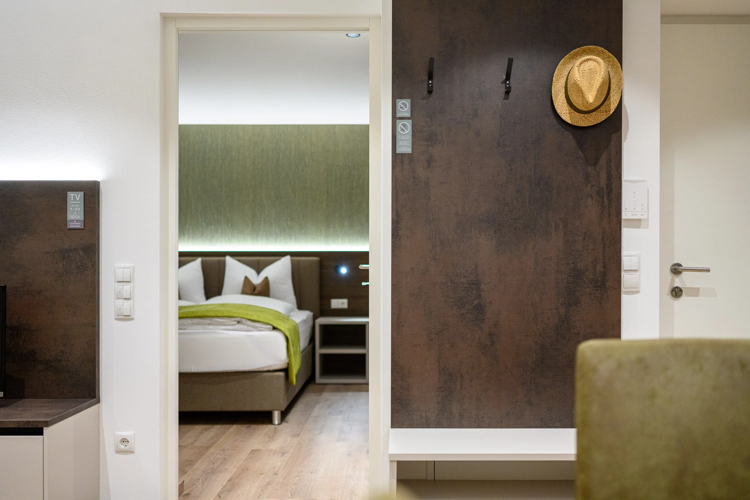 Hotel room with a bed, green bedding, pillows, a nightstand, and a green accent wall visible through an open doorway from a sitting area with a green chair.