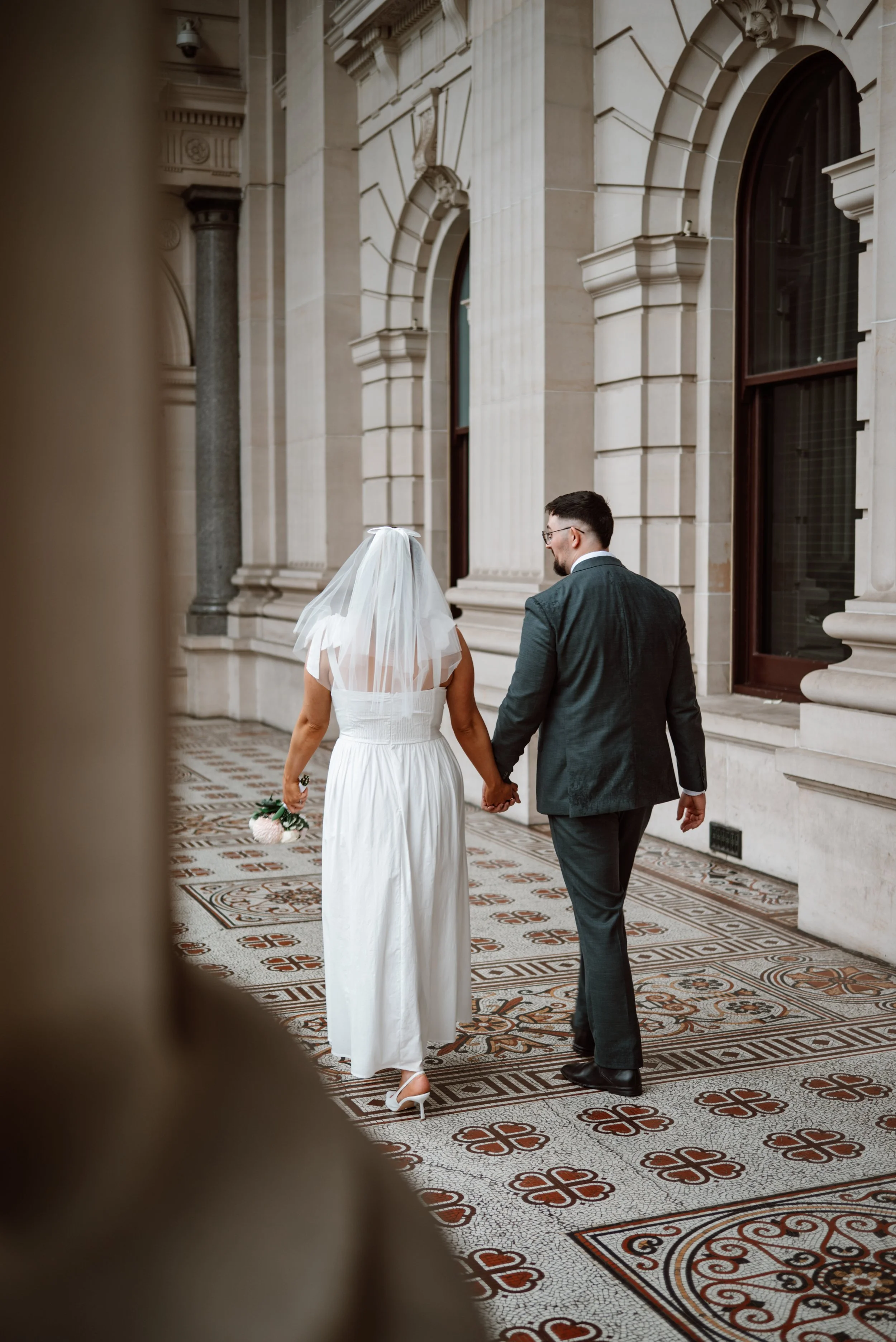 A bride and groom holding hands, walking away from the camera outside a grand, historic building with ornate stone architecture and large arched windows.