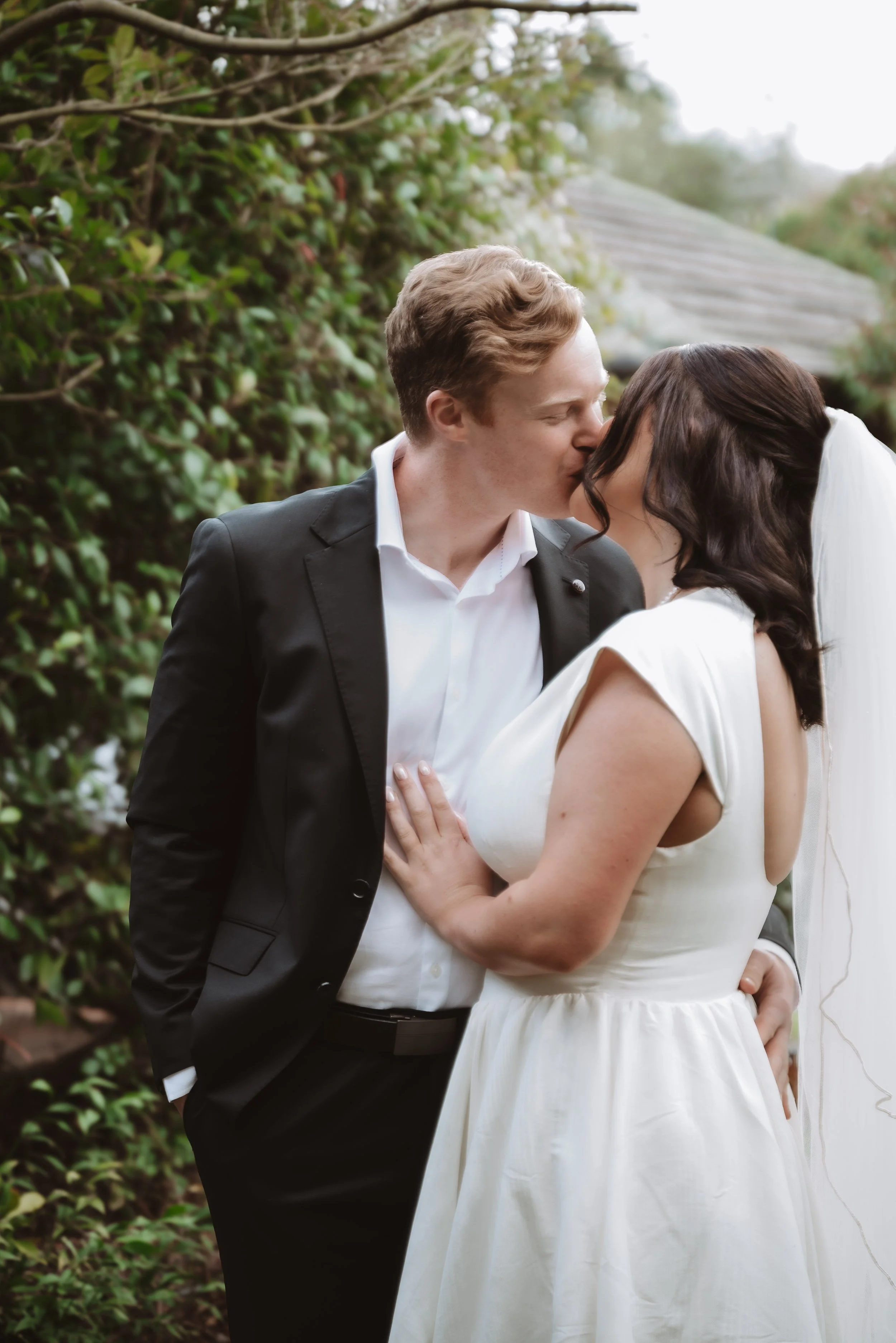 A couple in wedding attire sharing a kiss outdoors surrounded by greenery, with the groom in a black tuxedo and the bride in a white gown.