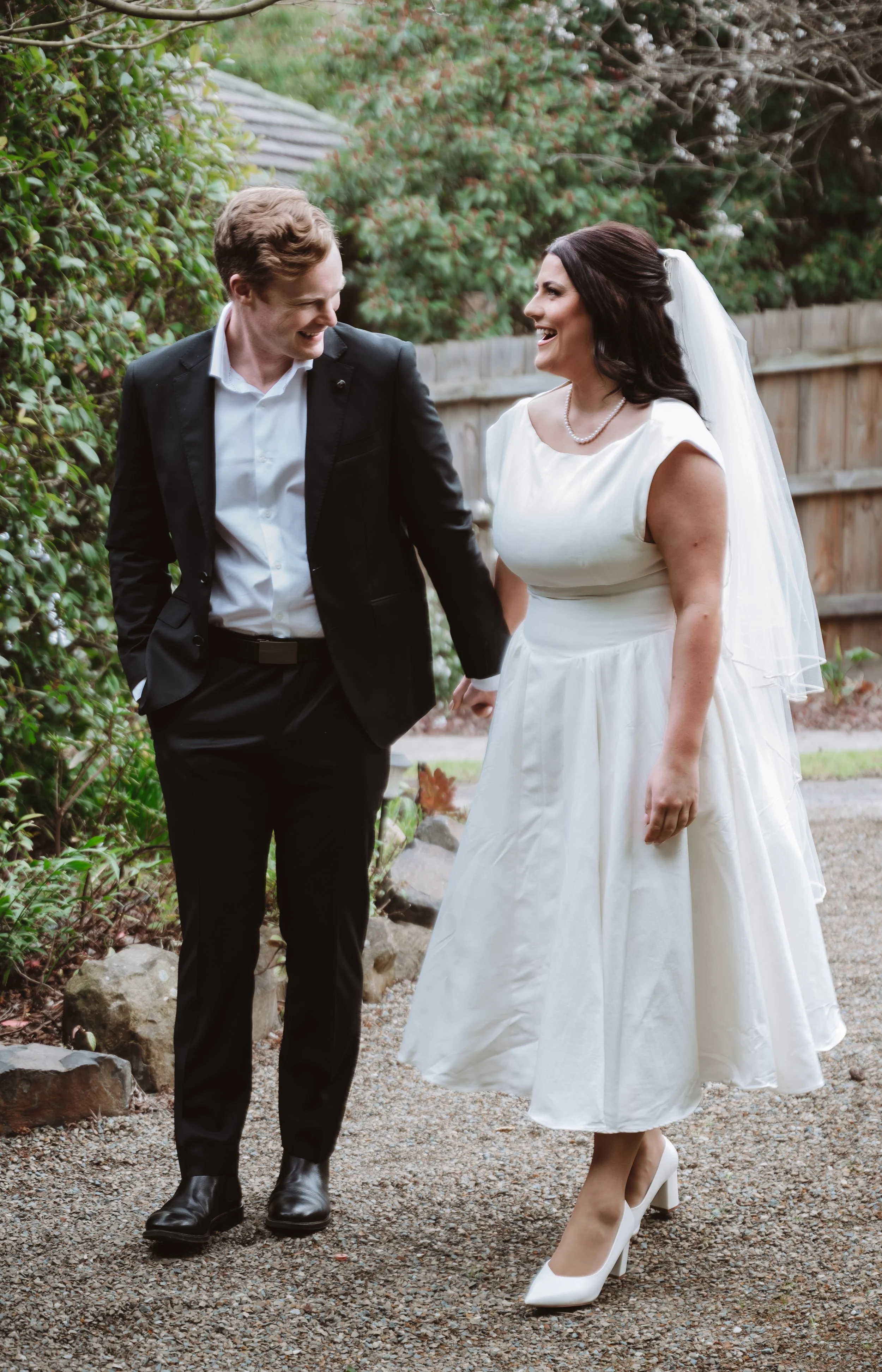 A bride and groom walking together outdoors, smiling, holding hands. The bride is wearing a white wedding dress, veil, and pearl necklace. The groom is in a black suit with a white shirt. They are surrounded by greenery and a wooden fence.