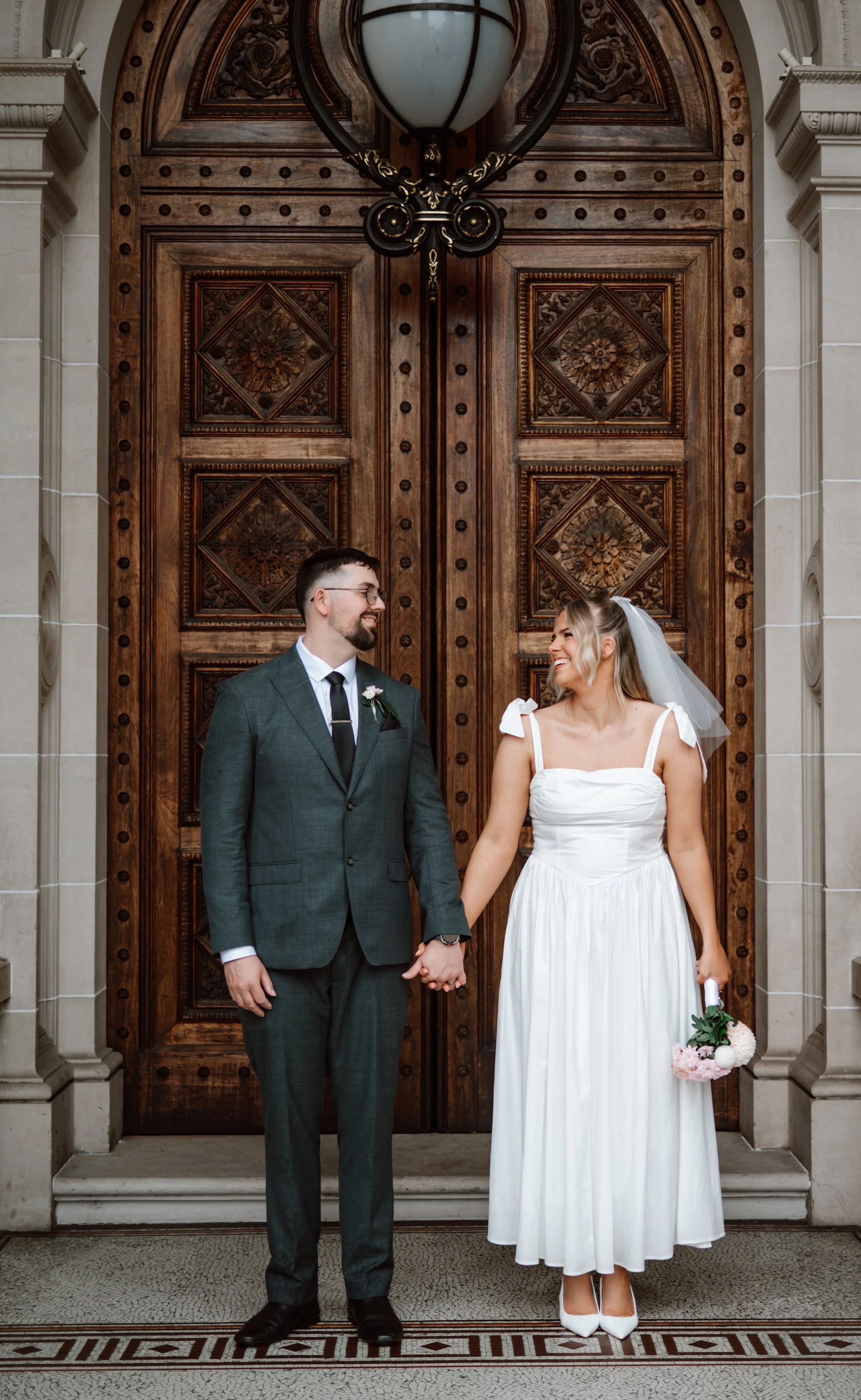 A bride and groom holding hands and smiling at each other in front of large wooden doors inside a church or courthouse, with the bride holding a bouquet of flowers.