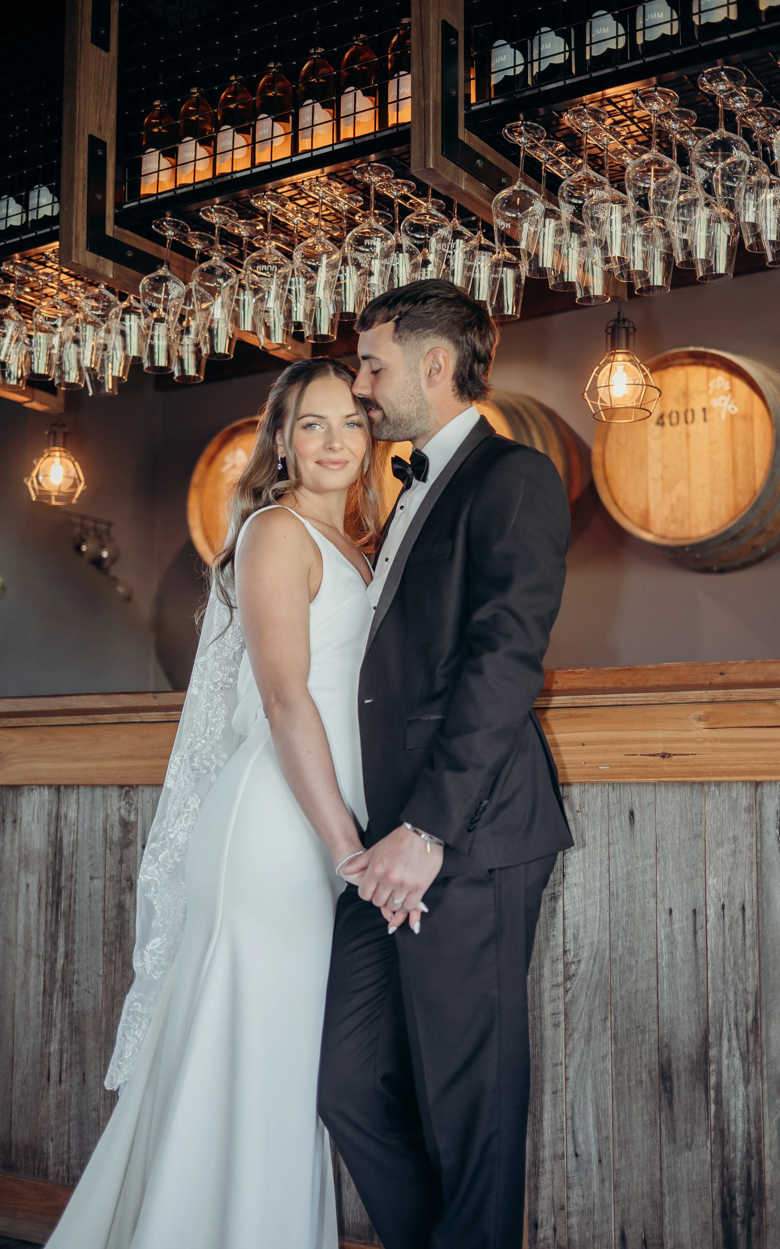 A bride and groom standing hand in hand outdoors, looking at a bison grazing near trees and a pond in a rural setting.