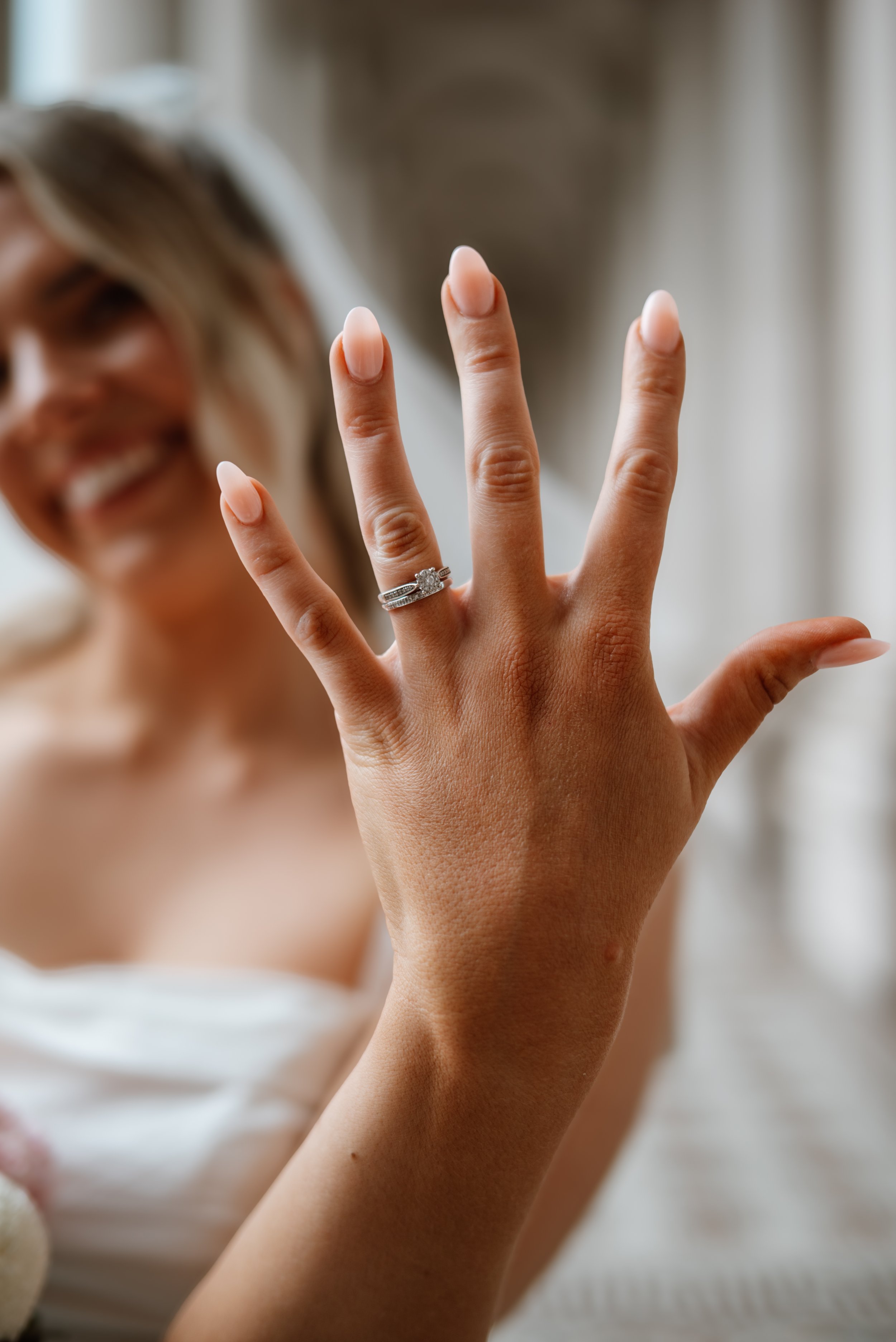 Close-up of a woman's hand showing a wedding ring, with her smiling face slightly blurred in the background.
