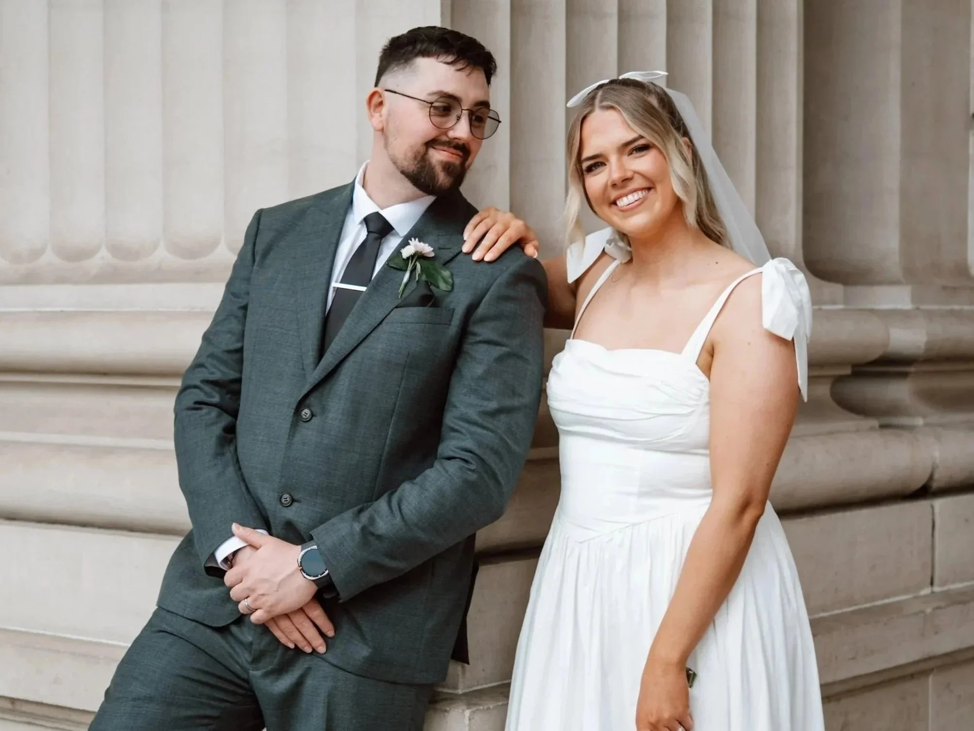 A smiling woman in a white dress with shoulder bows and a veil, and a man in a gray suit with a flower boutonniere, standing in front of a classical building with tall columns.