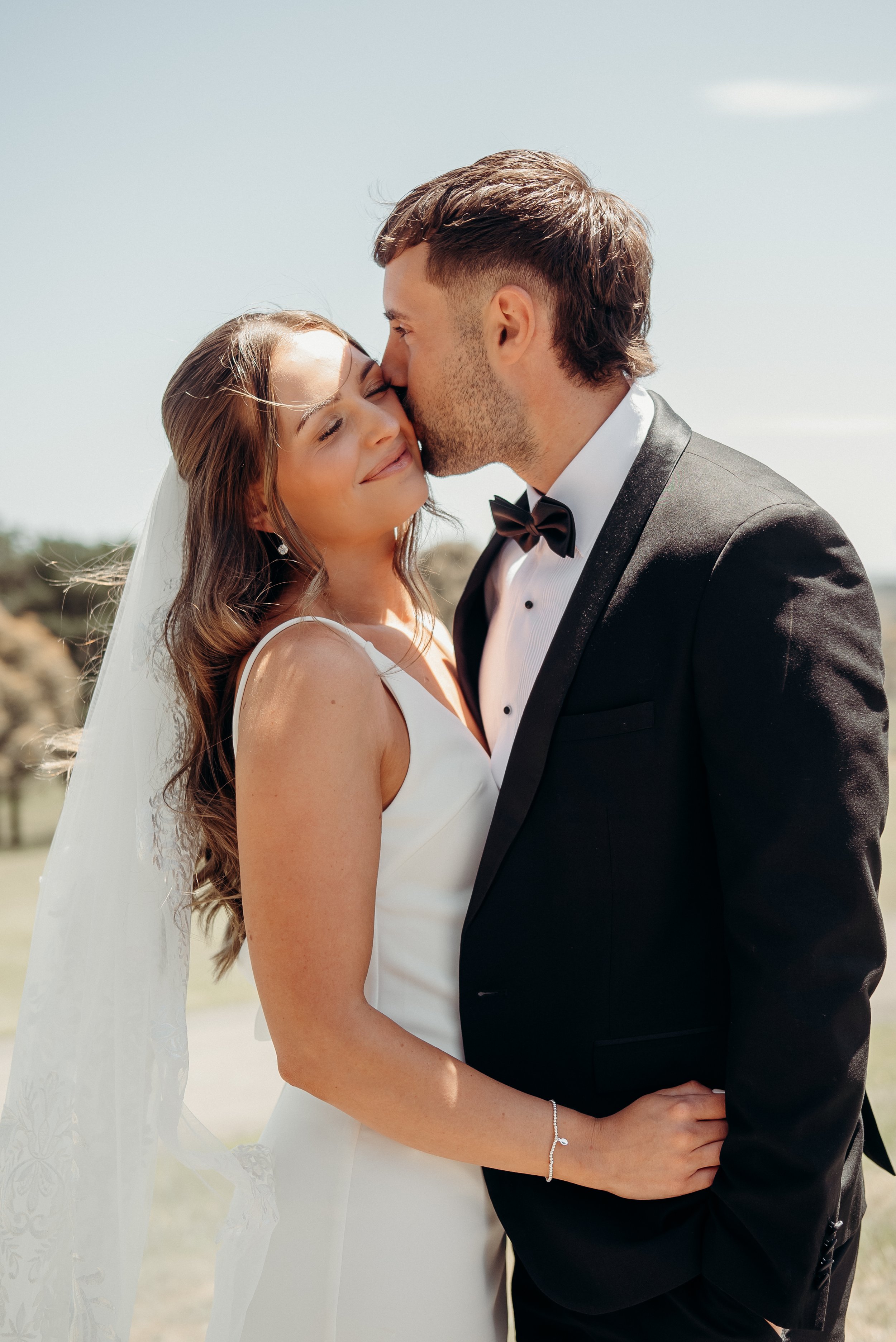 A bride and groom in wedding attire standing close together outdoors, with a large tree and a mountain in the background.