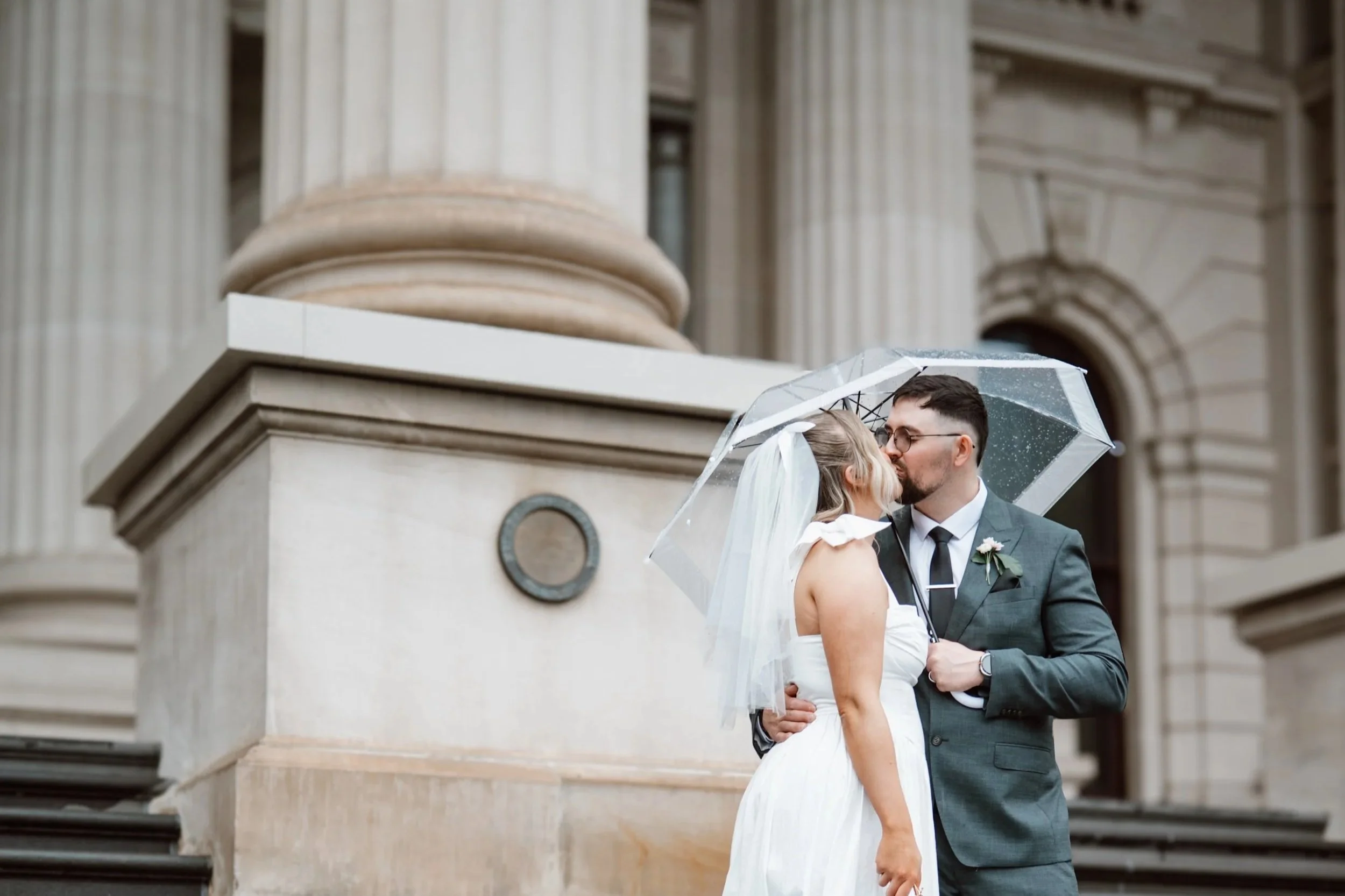 A newlywed couple sharing a kiss under a clear umbrella outside a historic building with large columns, after a wedding in the rain.