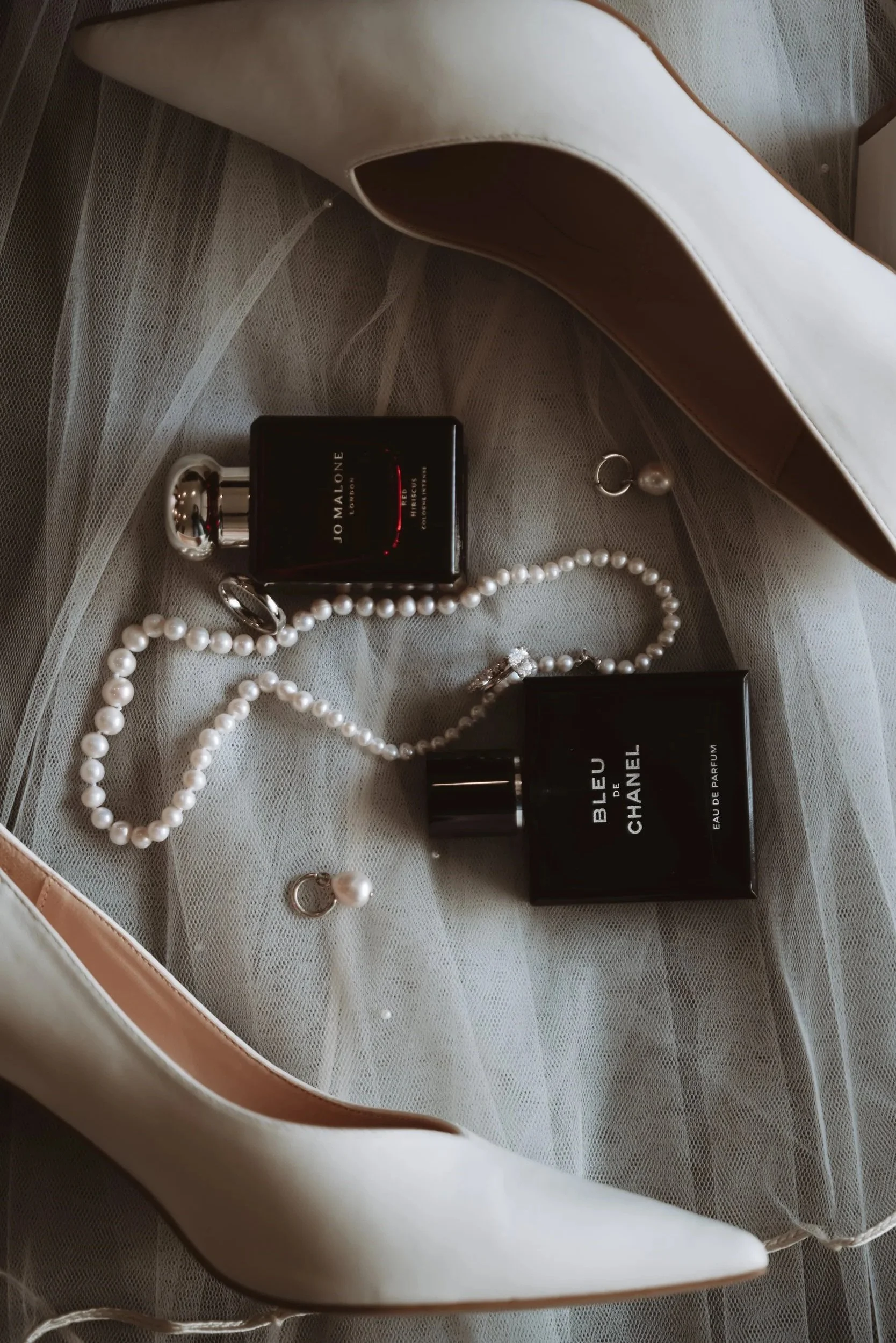 A white high-heeled shoe, two bottles of perfume, pearl jewelry, and a pair of pearl earrings arranged on a sheer fabric surface.