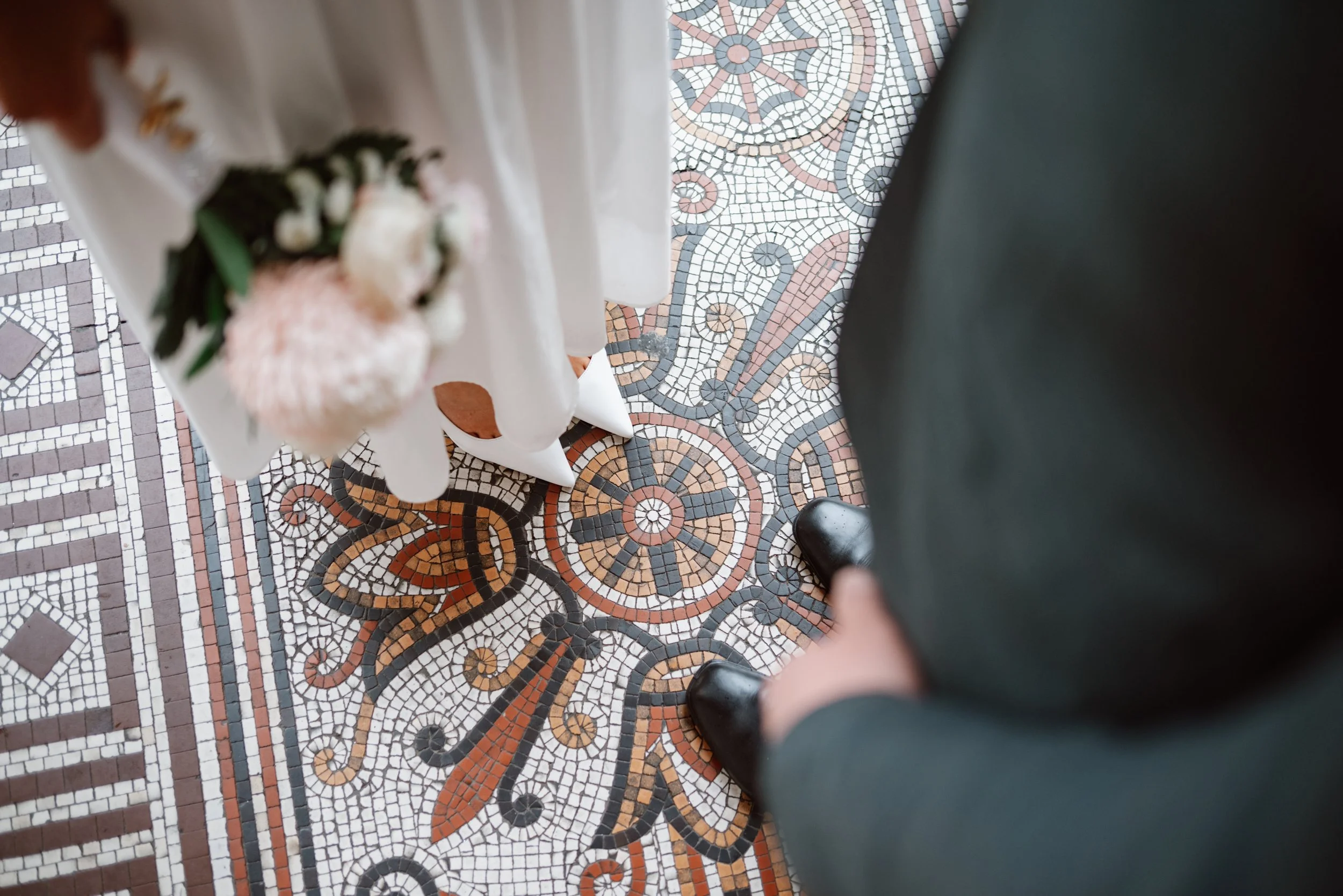 Looking down at someone's feet, wearing black shoes and dark pants, standing on an intricate mosaic tile floor with geometric and floral patterns in black, white, brown, and pink. Part of a person in a white dress holding a flower bouquet is visible to the left.