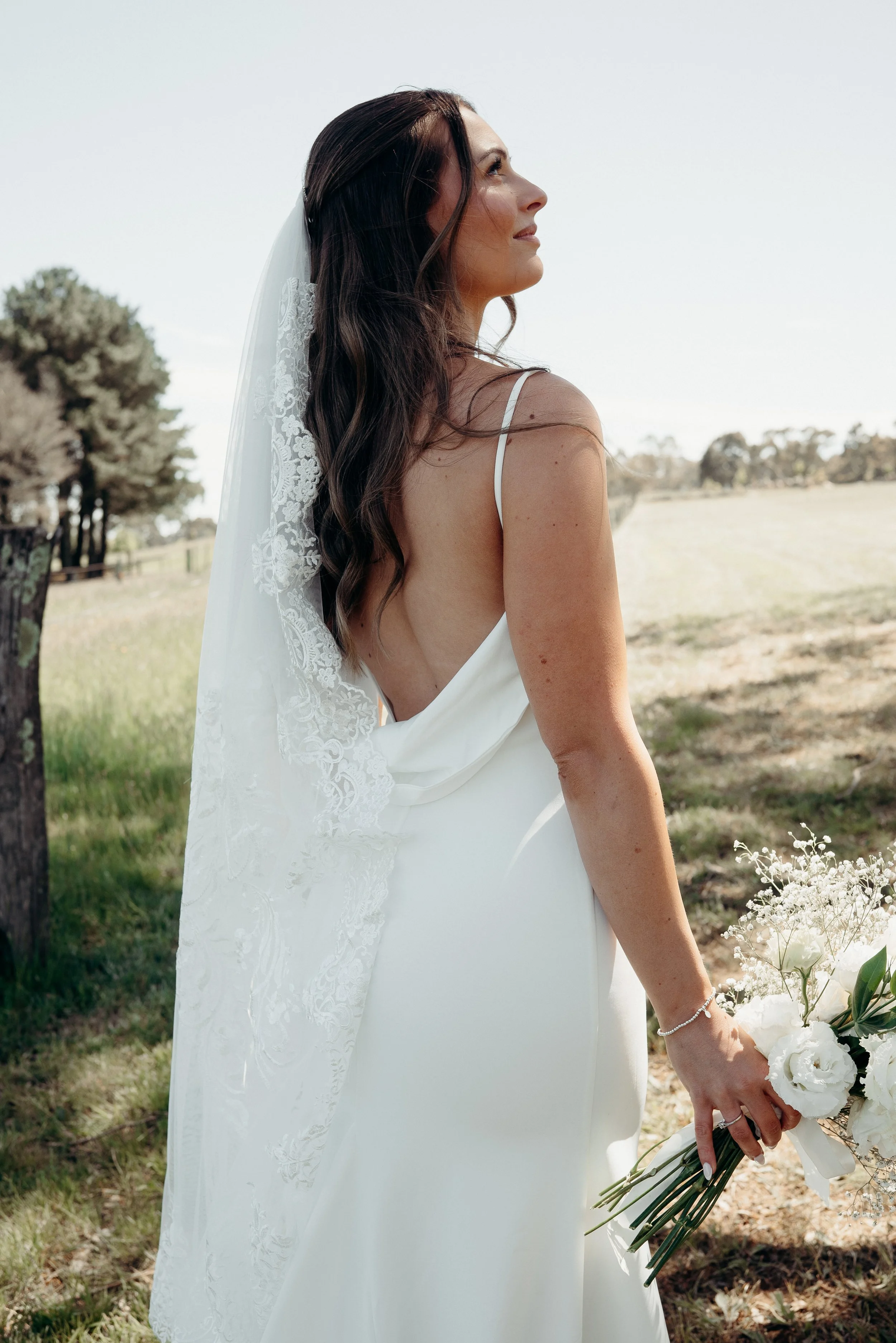 Bride in a white wedding dress with a lace veil holding a bouquet of white flowers outdoors, looking to the side.