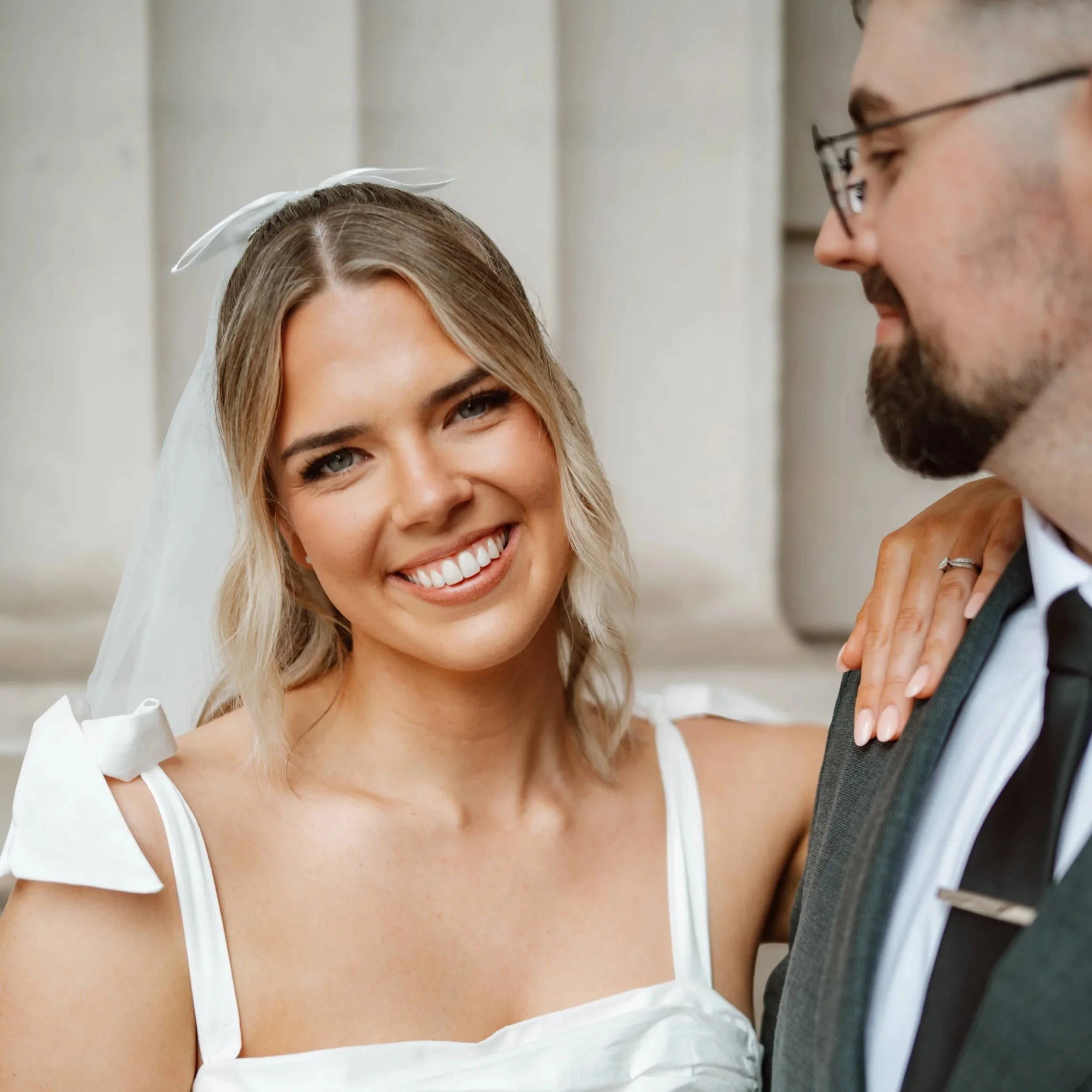 A bride with blonde hair and a big smile, wearing a white dress and veil, stands beside a groom in a suit. The bride has her hand on the groom's shoulder, showing a wedding ring. They are close together indoors.