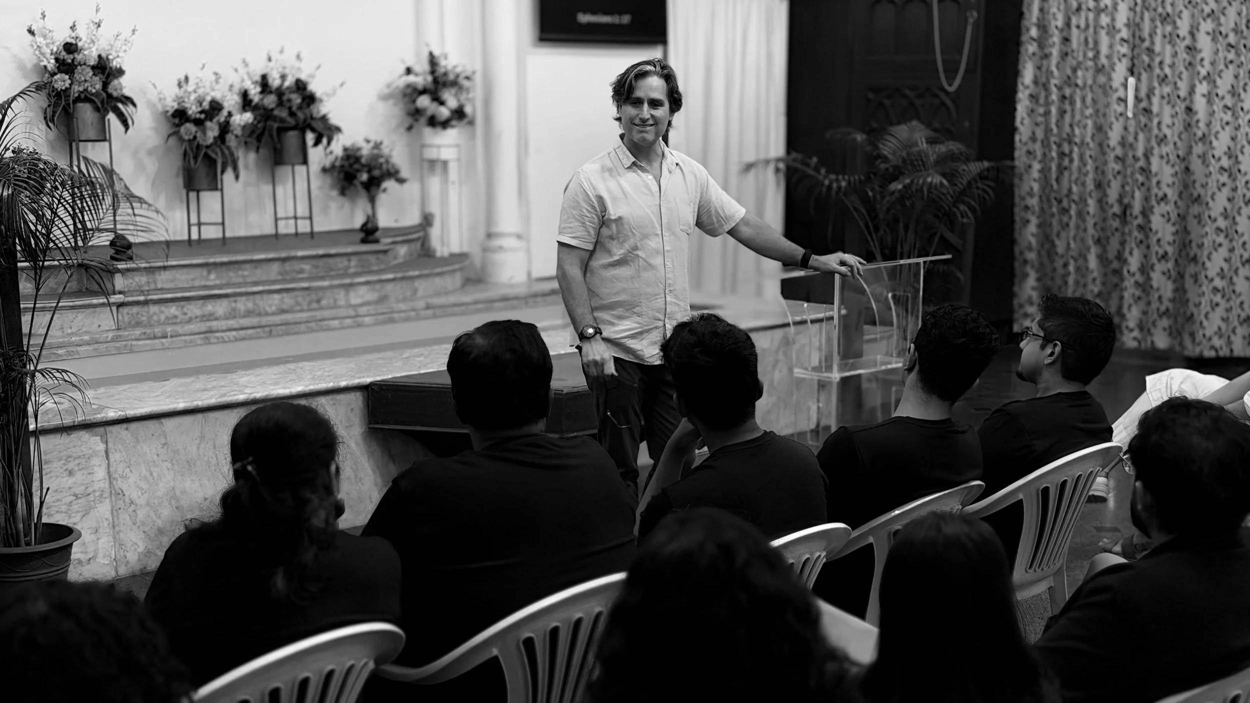 Ryan Waters tanding in front of an audience in a room with floral arrangements and curtains, presenting or speaking.