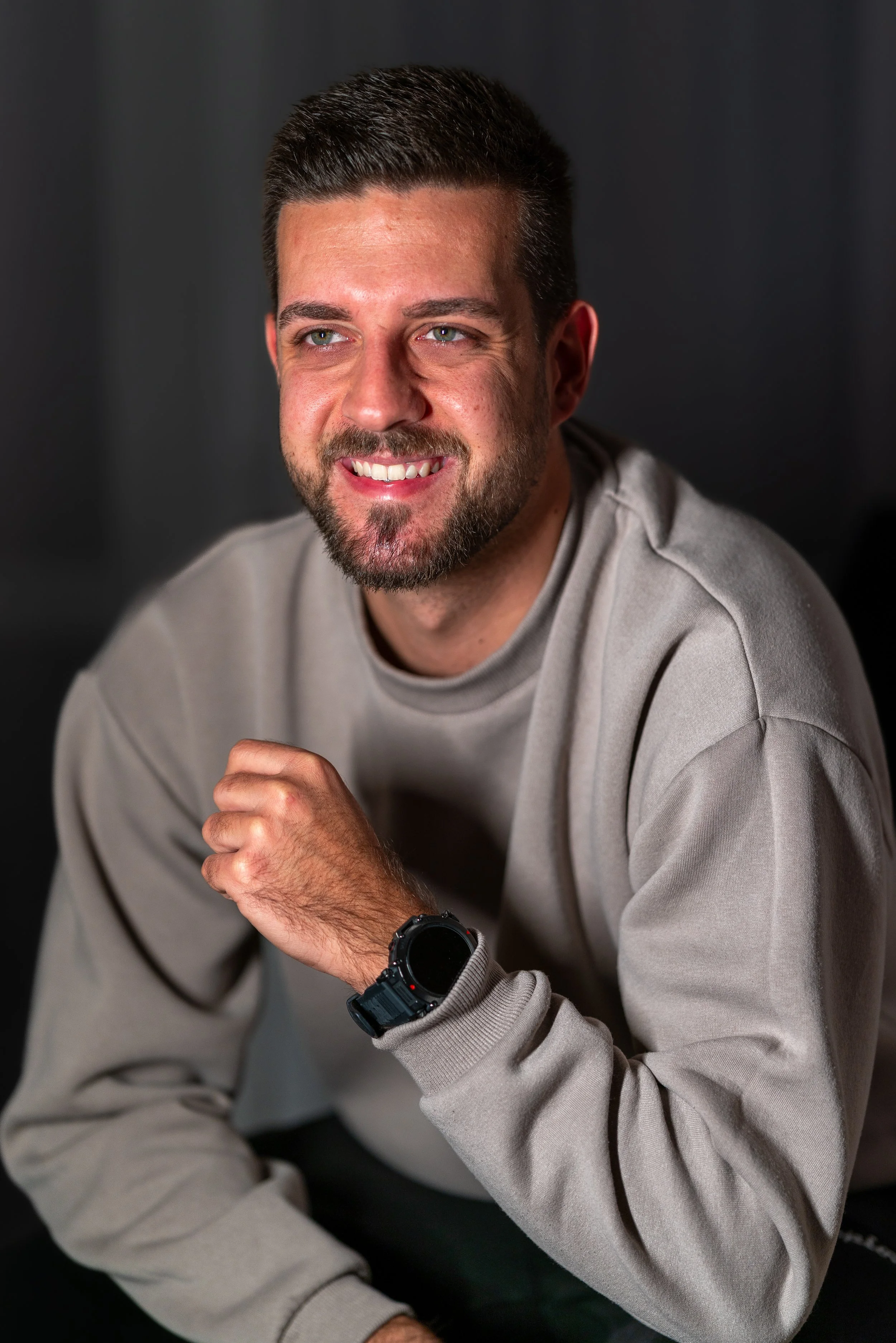 Ein lächelnder Mann mit dunklem Haar und Bart, trägt einen grauen Pullover und eine schwarze Smartwatch am Handgelenk, sitzt vor dunklem Hintergrund.