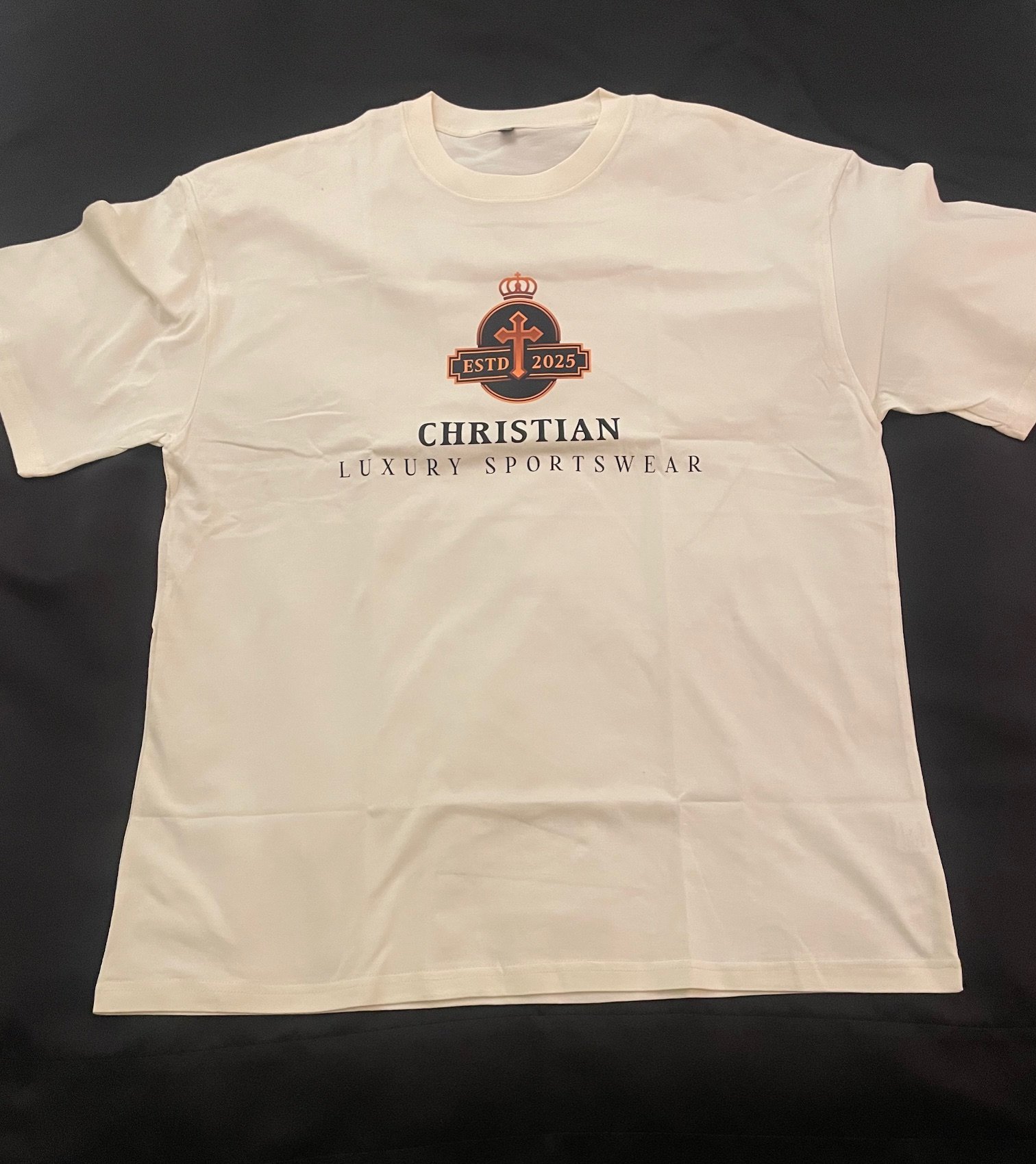 White T-shirt with a logo featuring a crown, cross, and the text 'ESTD 2025' in a circular design, and the words 'Christian Luxury Sportswear' printed below.