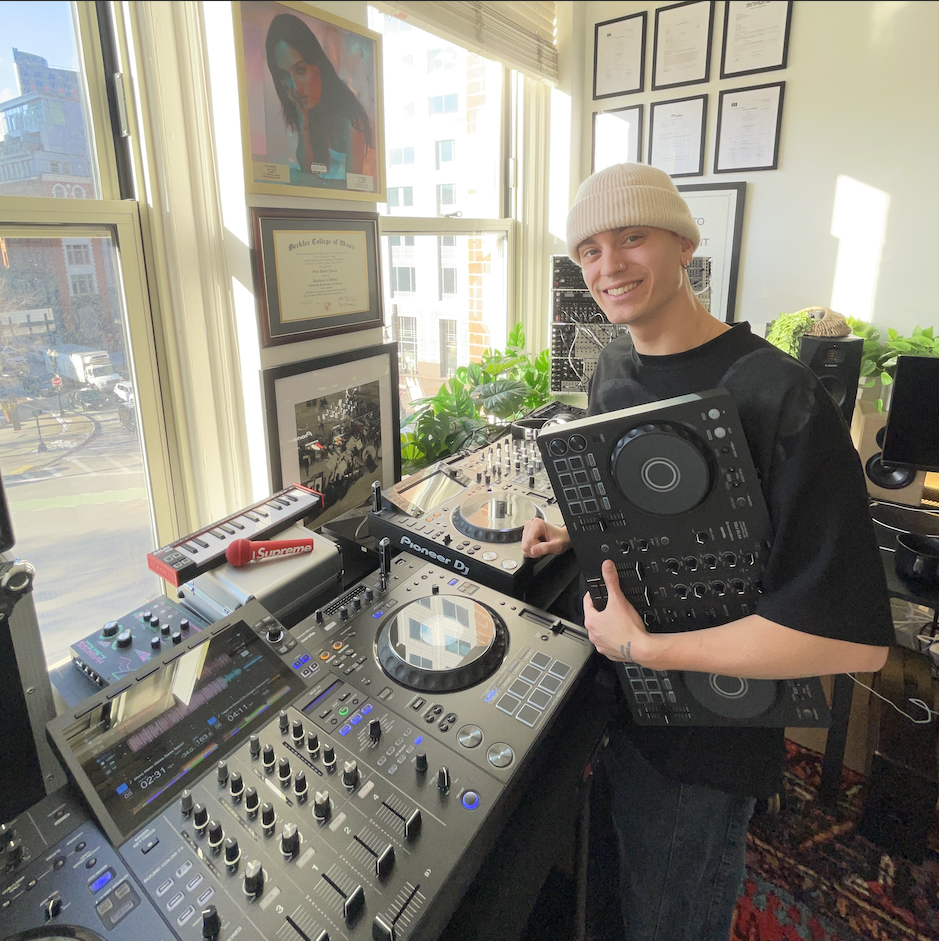 Young man wearing a white beanie and black t-shirt standing in a bright room with large windows, surrounded by audio equipment including a DJ mixer, turntables, and a controller, smiling at the camera.