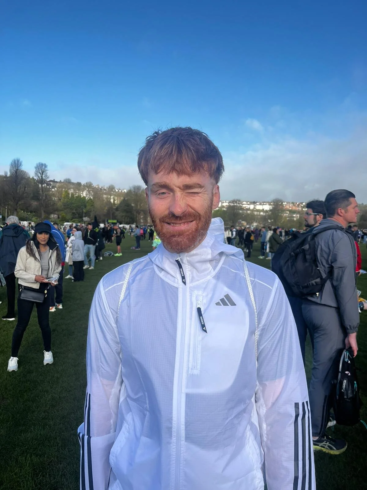 A man with a beard and short brown hair winking at the camera, wearing a white athletic jacket, outdoors in a grassy area with many people in the background, some holding backpacks, under a partly cloudy sky.